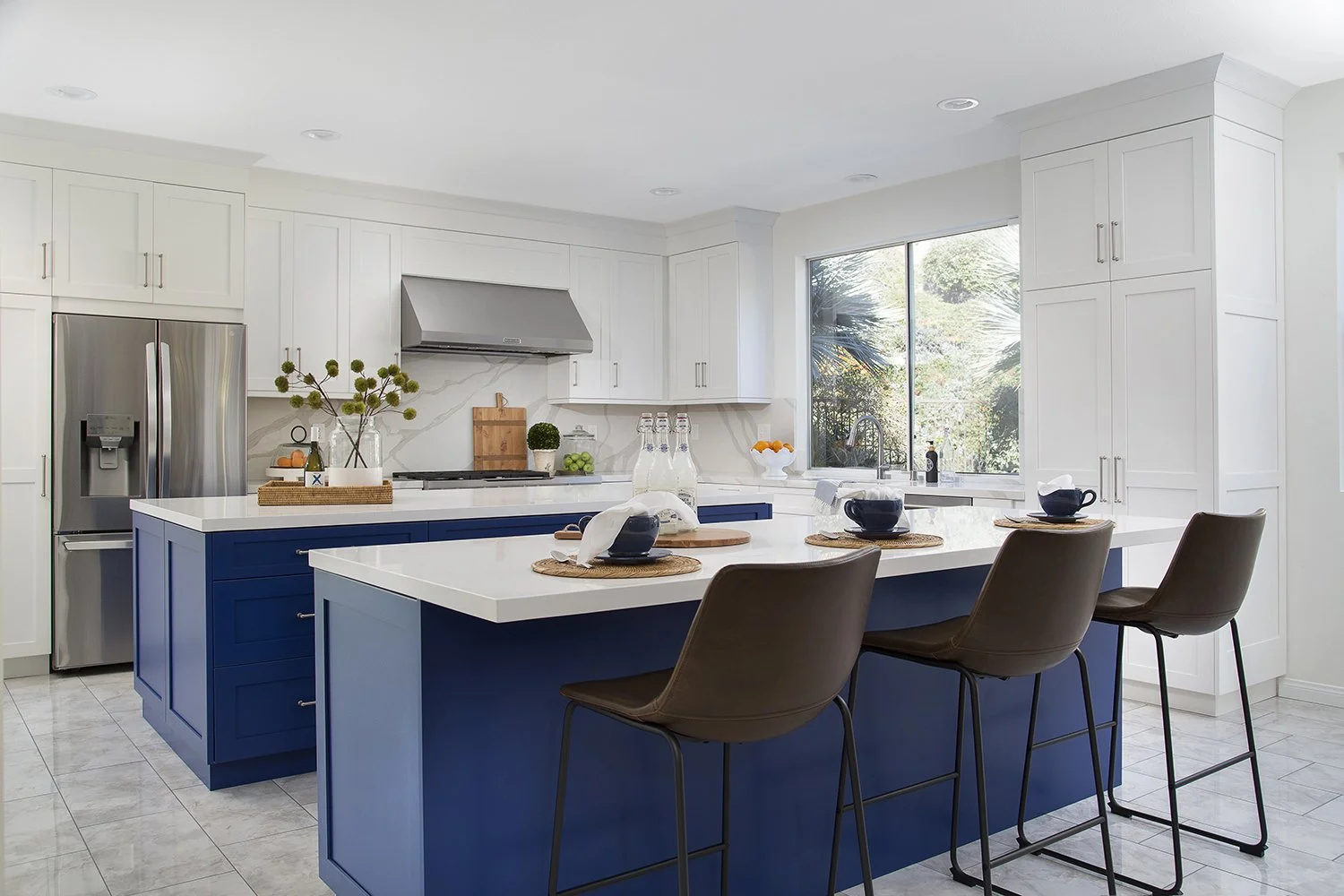 A modern kitchen features a large island with a blue base, white countertop, and three brown bar stools. The kitchen has white cabinets, a stainless steel refrigerator, and a large window letting in natural light. Decor includes plants and various ki