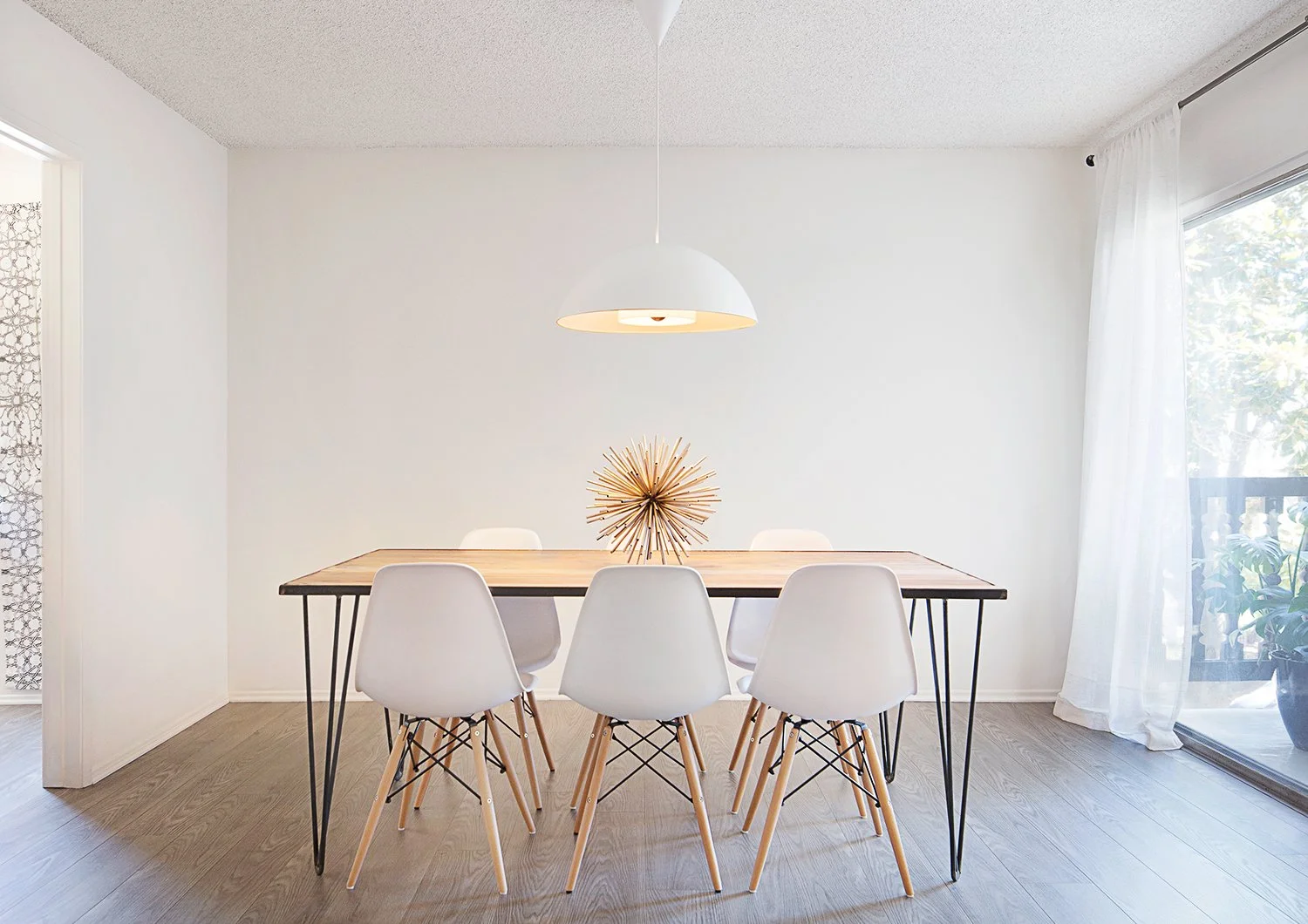 Minimalist dining room with a wooden table, five white chairs, and a sunburst decor piece as a centerpiece. A modern pendant light hangs above the table. The room features light-colored walls, hardwood floors, and a large window with sheer curtains.
