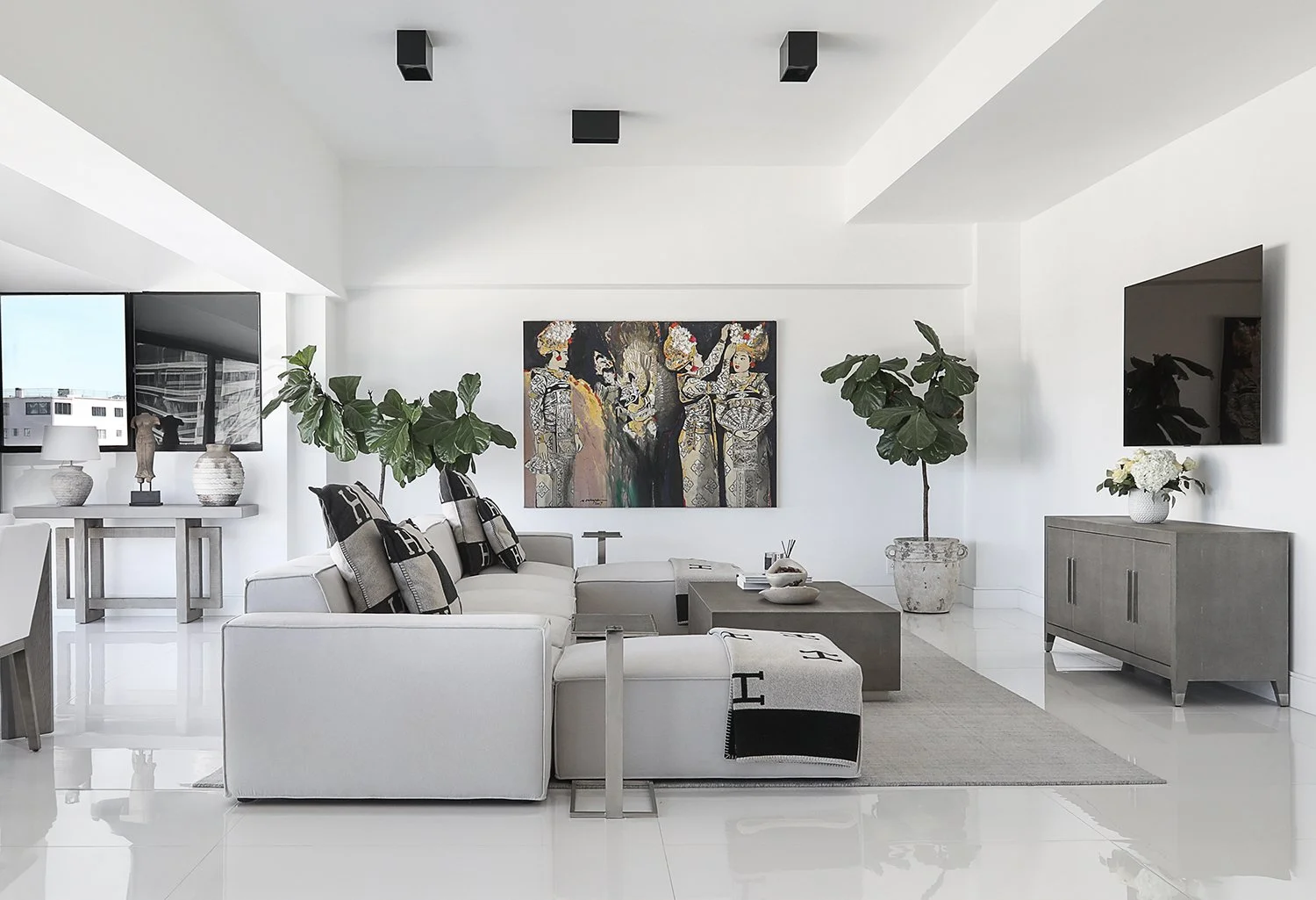 A modern, minimalist living room with white walls and floor, featuring a gray sectional sofa with cushions, a rectangular coffee table, a large abstract painting on the wall, two potted plants, a gray sideboard with a flat-screen TV, and a dining are