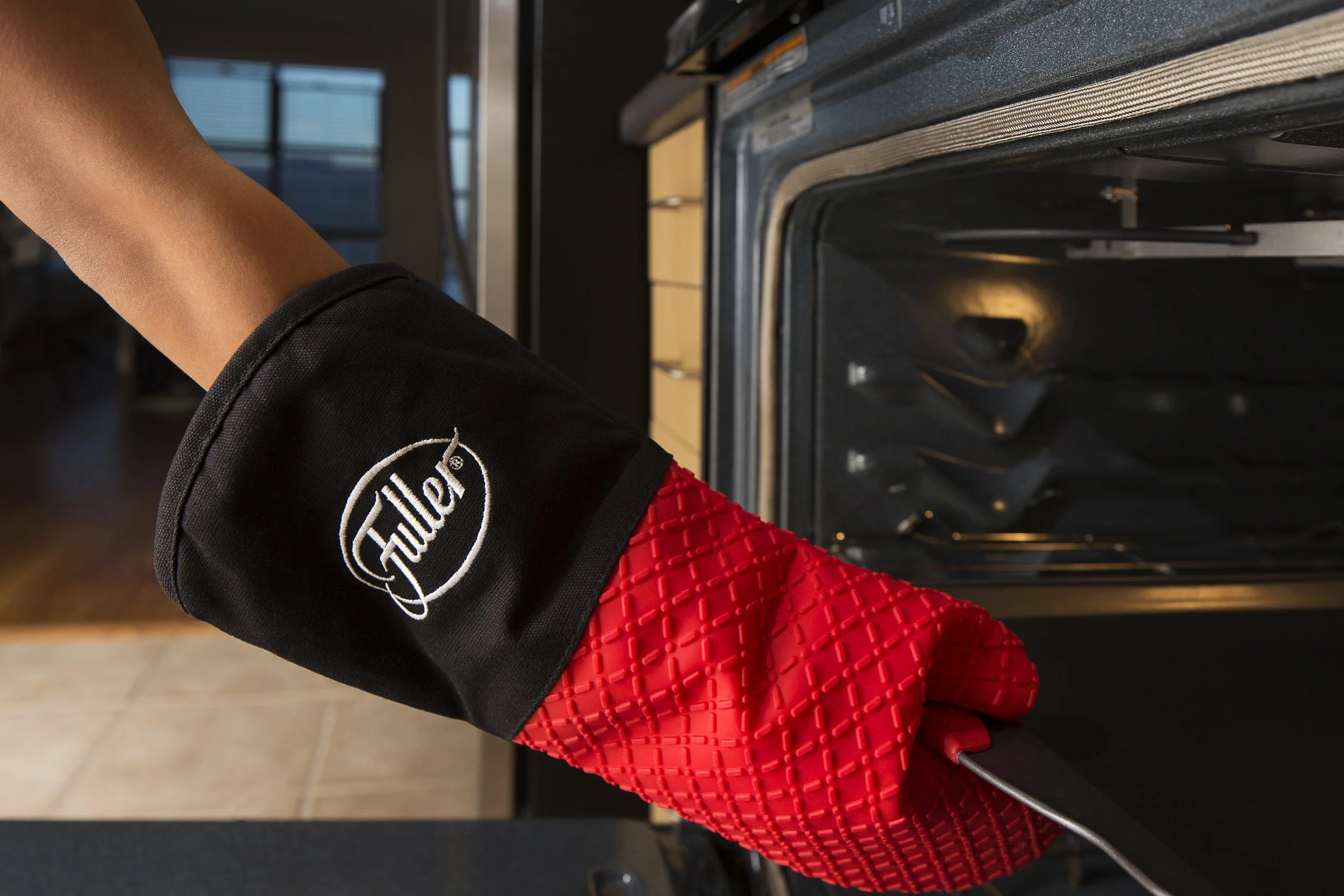 A person wearing a red and black oven mitt with "Fuller" written on it is placing or removing a tray from an open oven in a kitchen.