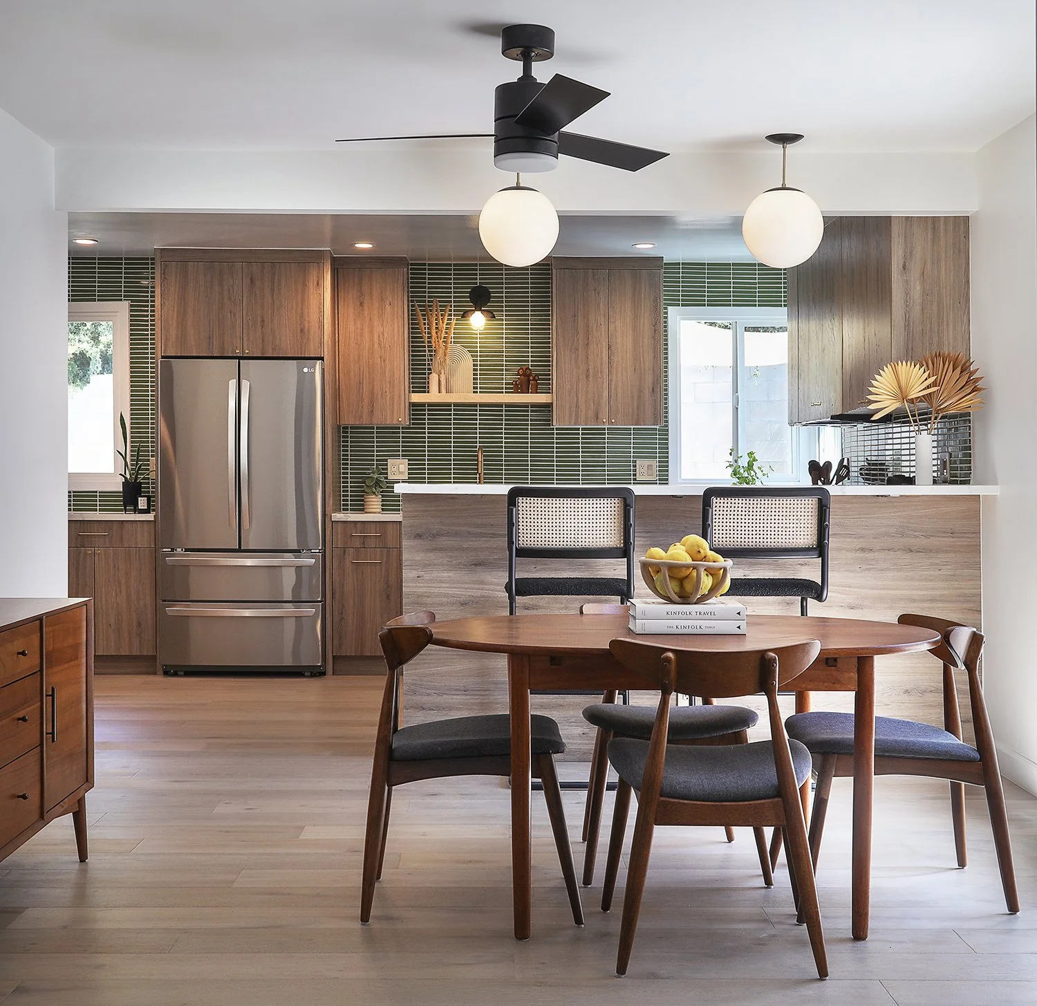 A modern kitchen and dining area with wooden cabinets, stainless steel refrigerator, and green tile backsplash. The dining table has four chairs and a bowl of lemons. There's a ceiling fan and two spherical hanging lights. The floor is light-colored 