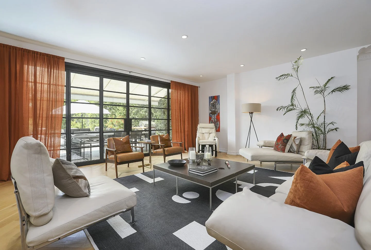 Modern living room with large windows, orange curtains, neutral sofas with orange and gray pillows, a black-and-white geometric rug, potted plant, and a coffee table. Patio and greenery are visible outside.