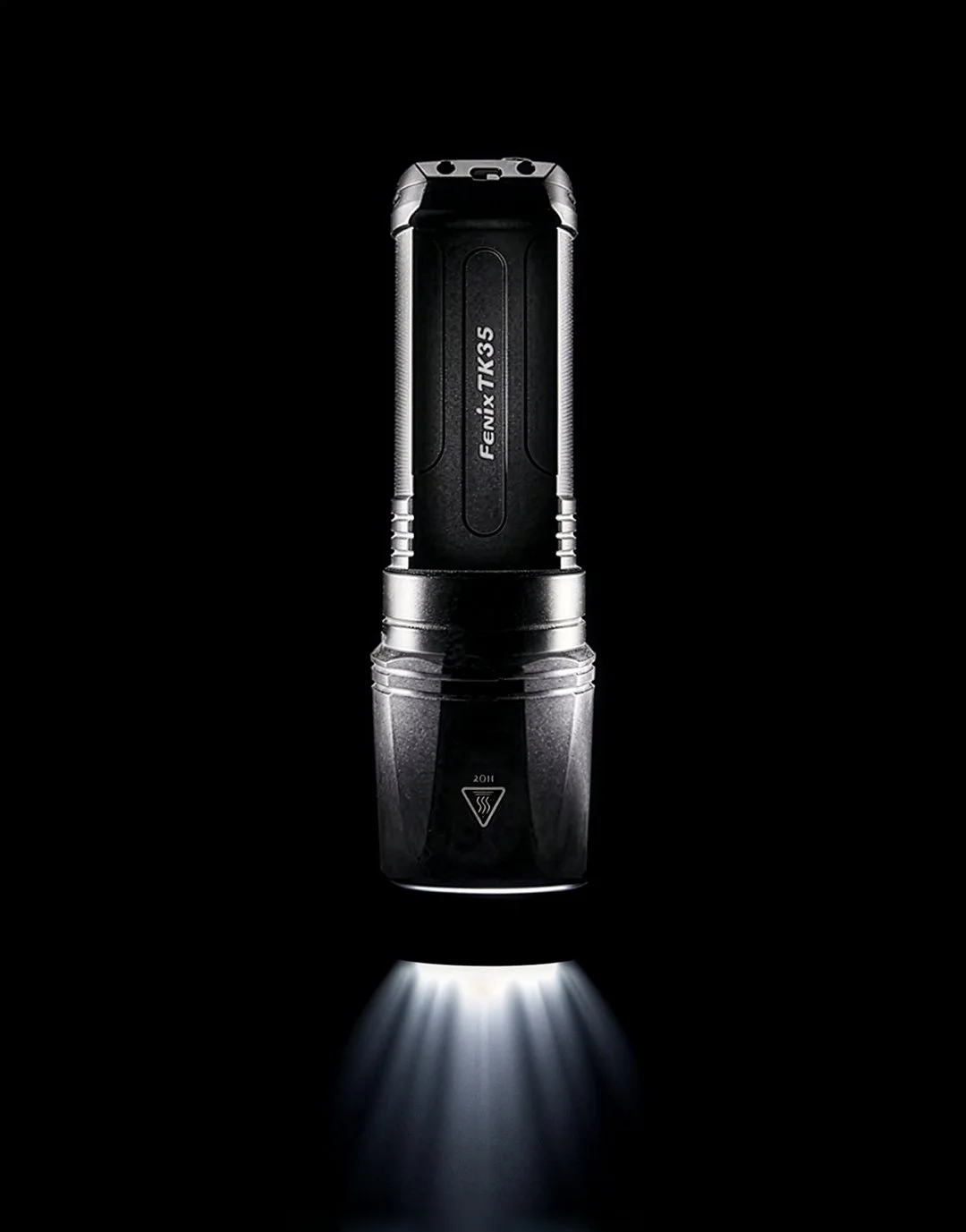 A black Fenix TK35 flashlight is standing upright in the dark, emitting a bright white beam of light downward, with light rays visible on the surface below.