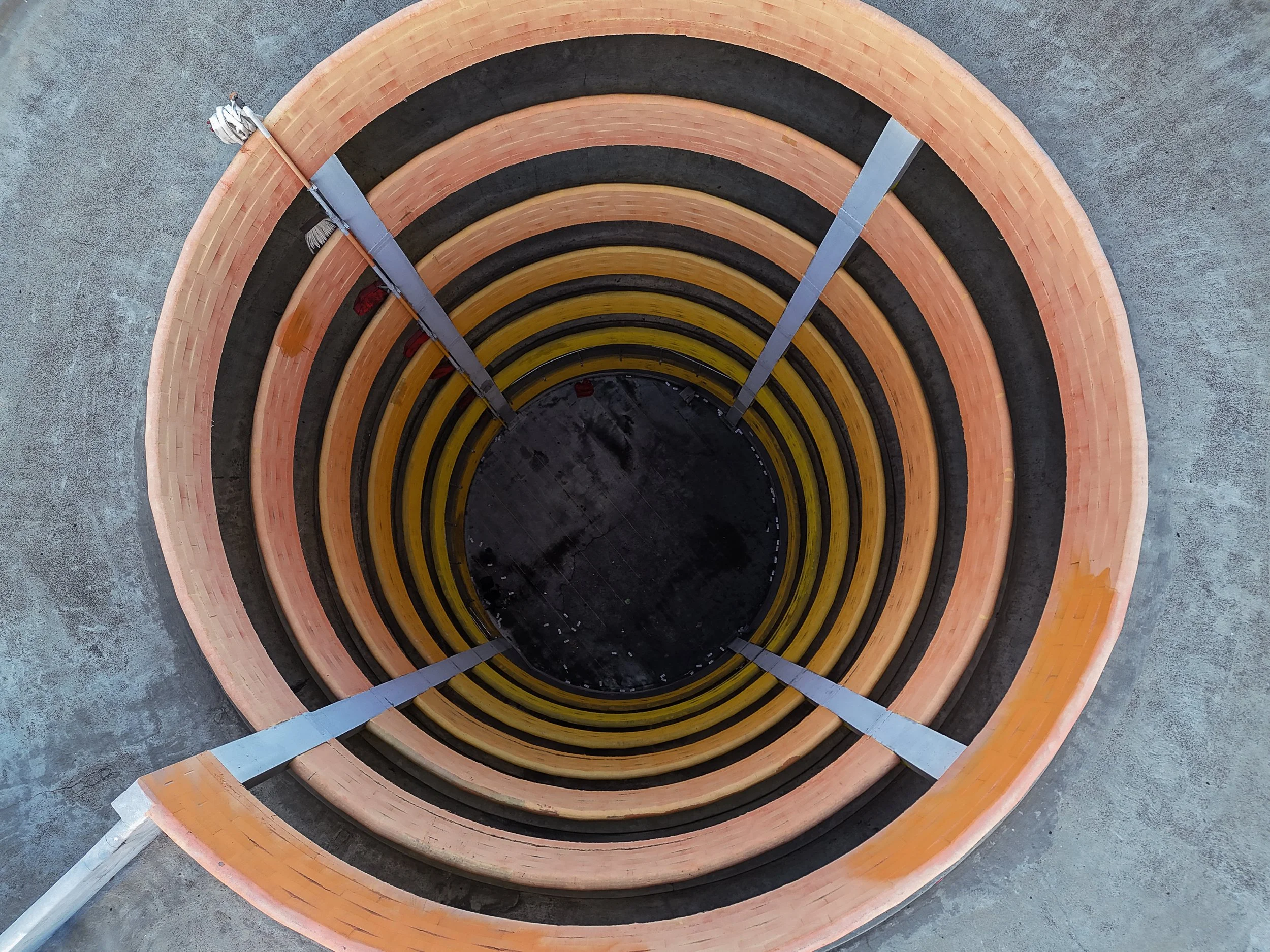 Aerial view of a circular, layered structure with alternating black and brown rings, resembling a vortex or tunnel. The outer layer is light brown, and the inner area appears dark, with a smooth concrete surface surrounding the structure.