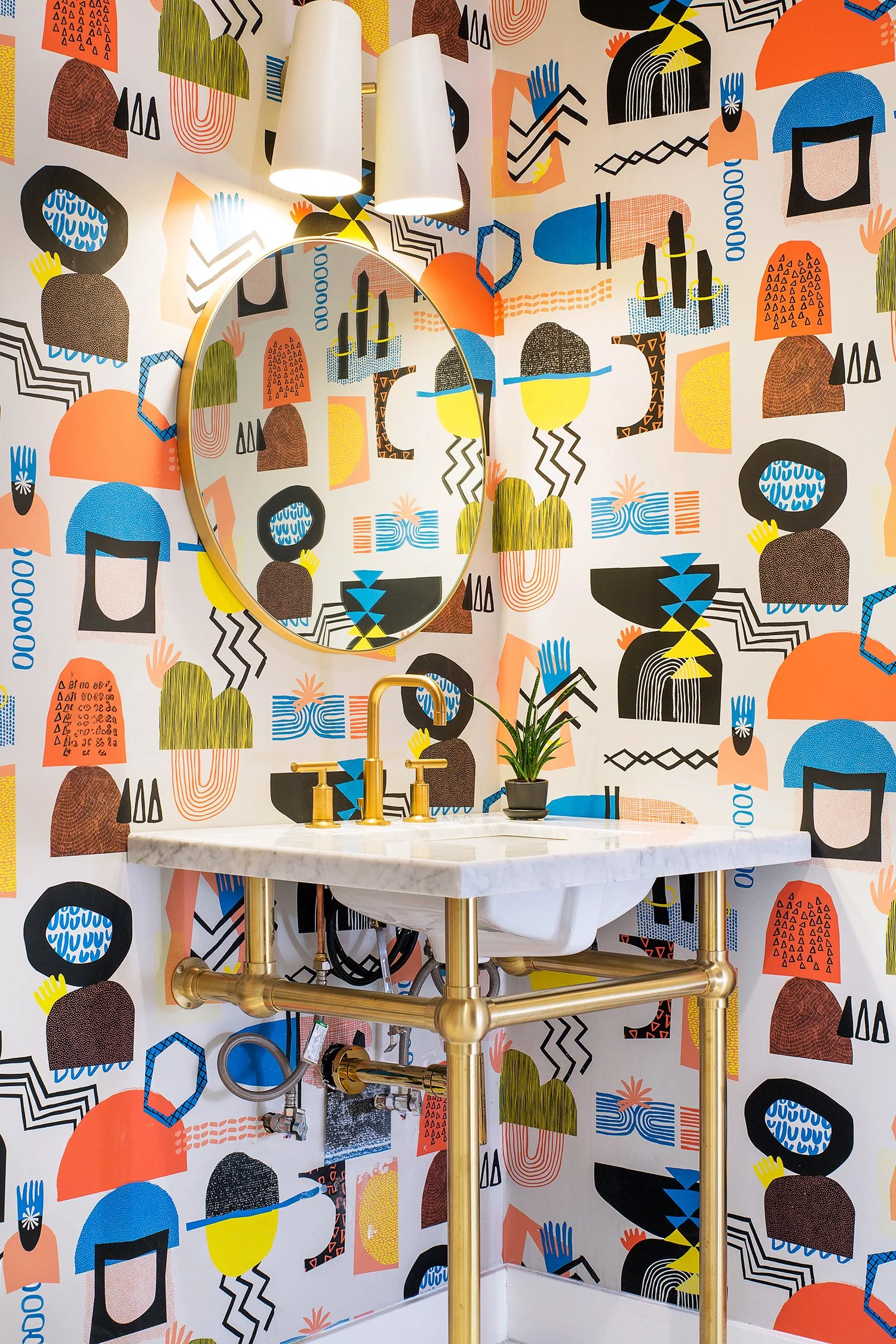 A small bathroom with a vibrant, abstract-patterned wallpaper in various colors. It features a marble sink with a brass fixture, a round mirror with a brass frame, two wall-mounted light fixtures, and a small, potted plant on the sink counter.