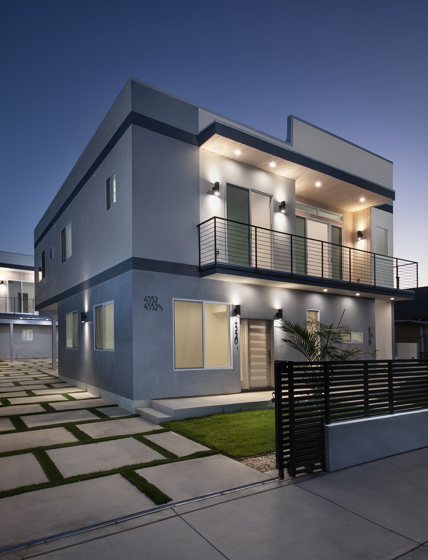 Modern two-story house with clean, geometric design, featuring large windows, a balcony with sleek black railing, and ambient lighting. The exterior walls are gray with minimalistic design elements. A paved driveway with grass inlays leads to the pro