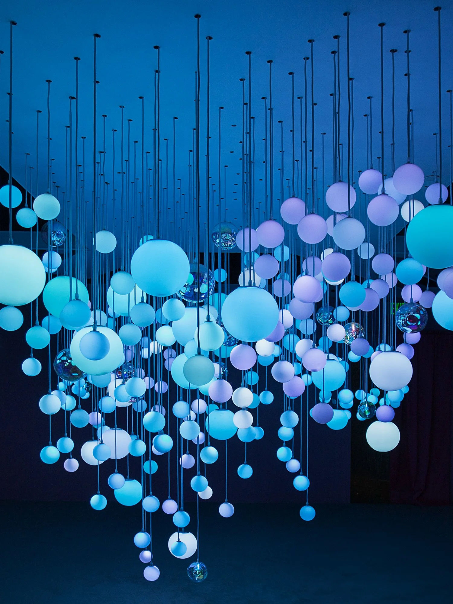 An artistic installation at MODS of numerous glowing orbs of various sizes suspended from the ceiling by thin wires. The orbs emit blue and purple light, creating a surreal, ethereal atmosphere. The background is dark, enhancing the orbs' luminescenc