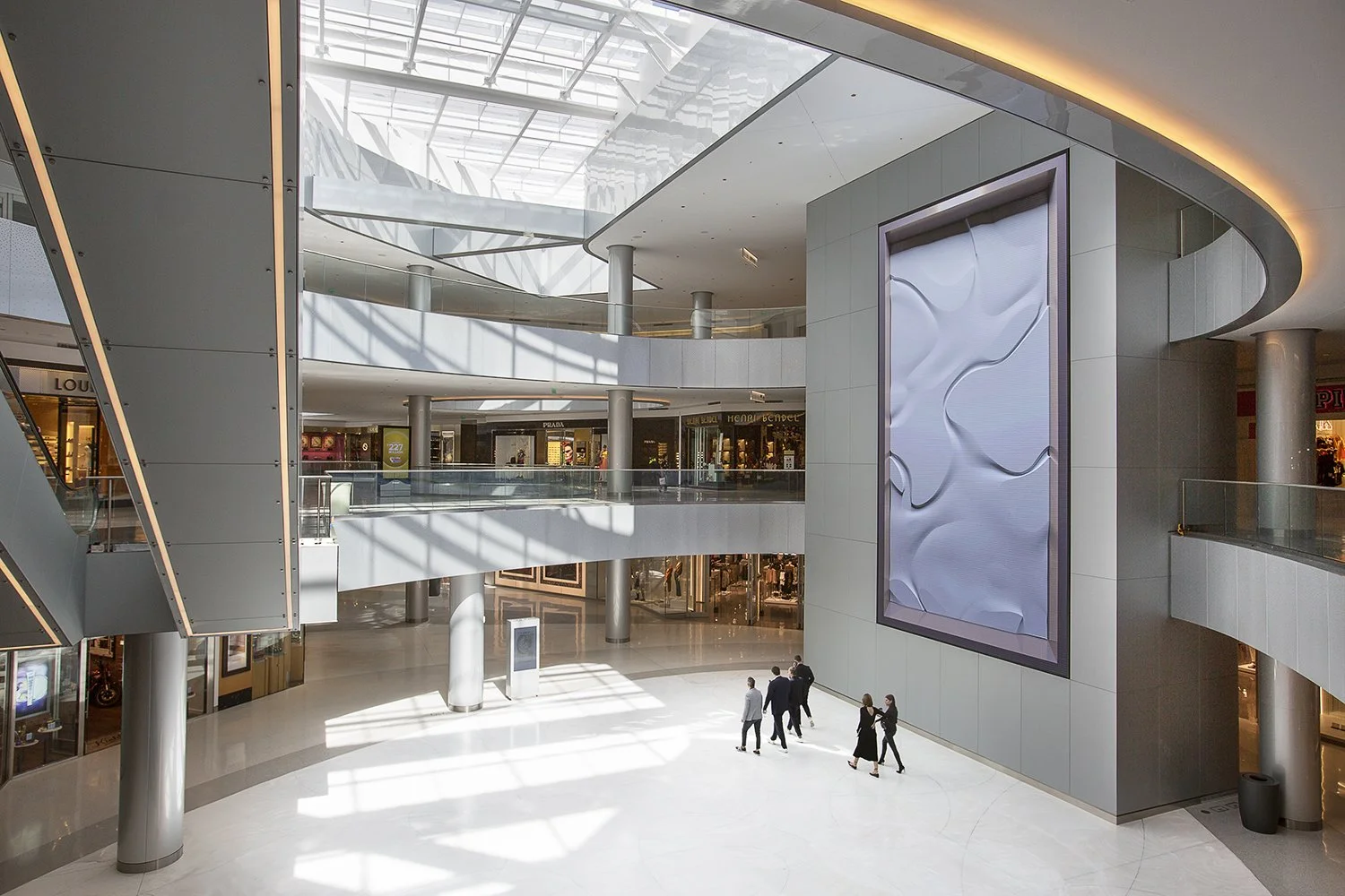 A spacious, modern shopping mall interior at the Beverly Center with a high glass ceiling and sleek design. People walk along a polished floor surrounded by stores. A large abstract art by Refik Anadol is prominently displayed on the wall.
