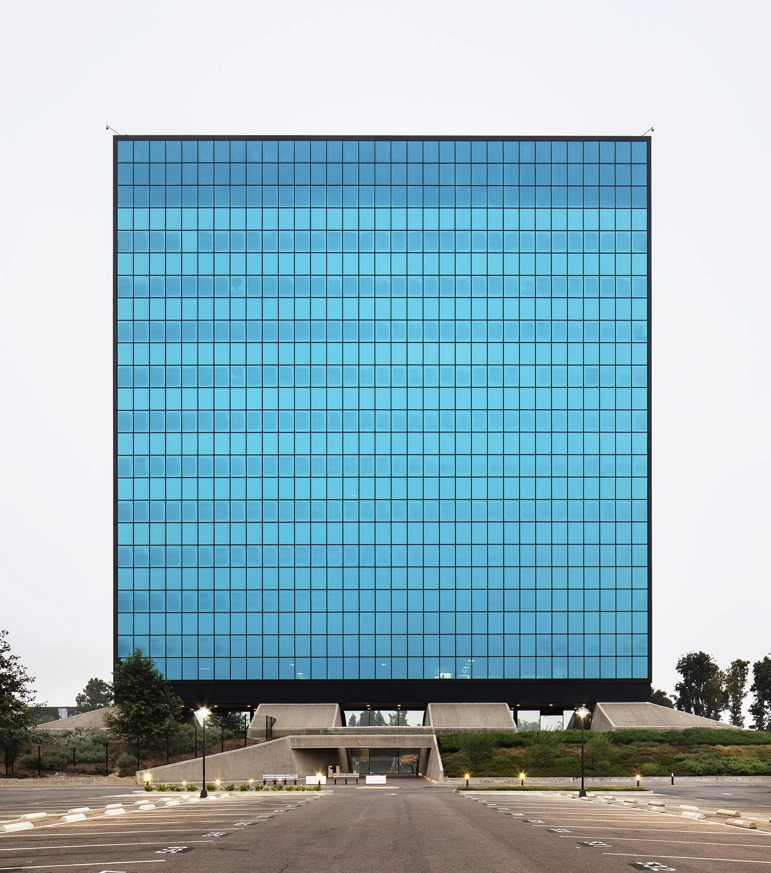 A large, modern cubic office building in Alhambra, CA, with reflective blue-tinted windows sits on a raised platform. The surrounding area is a mostly empty parking lot with sparse trees. The building is now owned by the Los Angeles of Department and