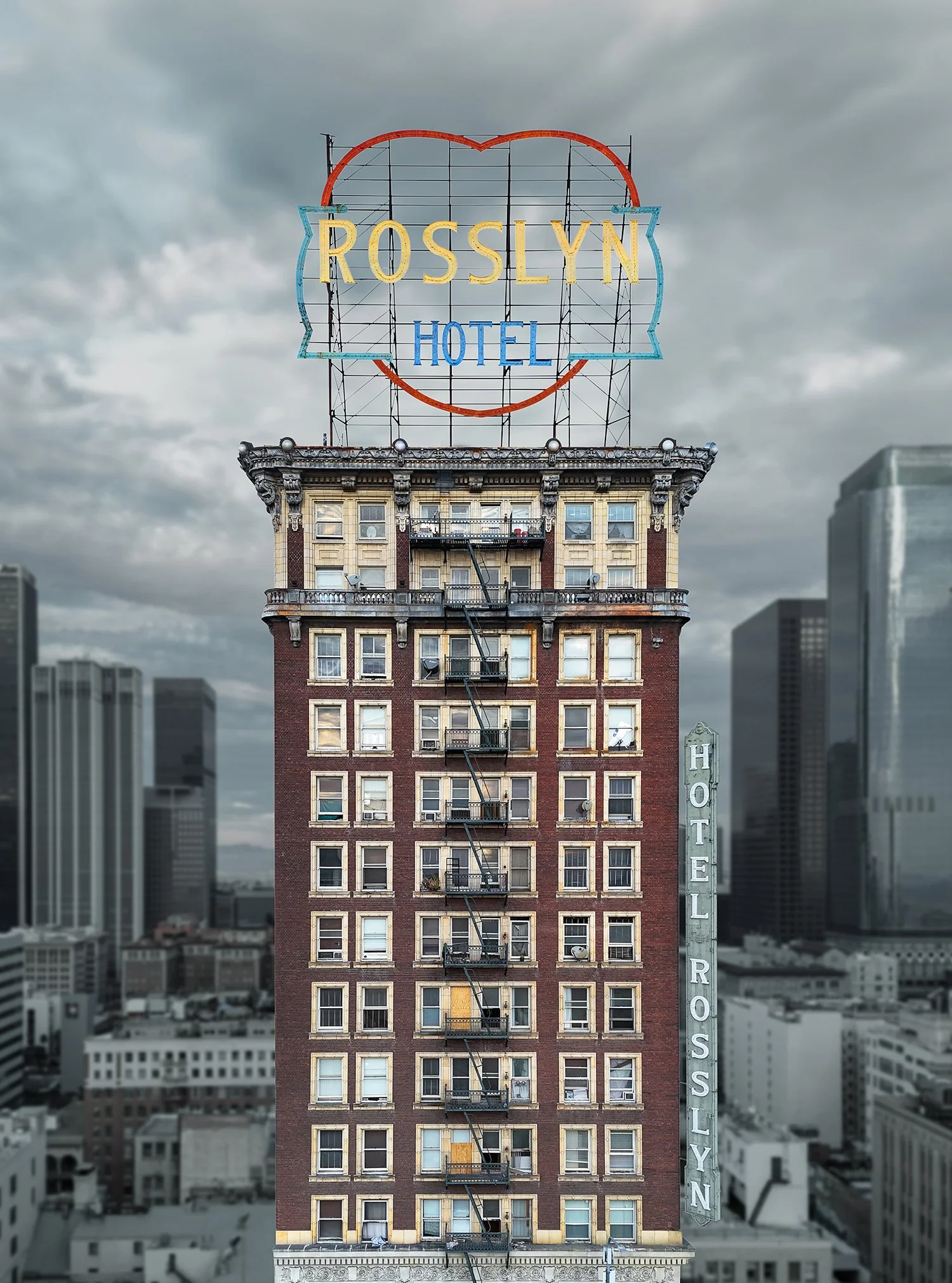 A tall, historic brick building located in Los Angeles, named the Rosslyn Hotel is shown. It has an old-fashioned neon sign on the roof depicting a heart shape around the name "Rosslyn." The sky is overcast, and other skyscrapers are visible in the b
