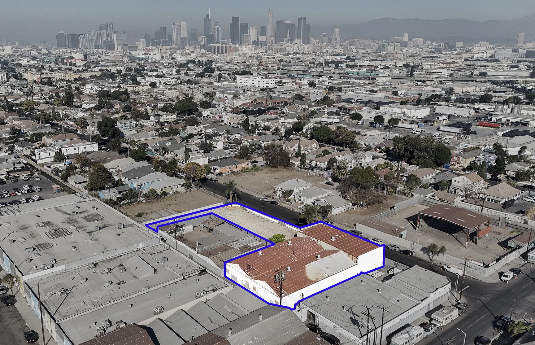 Aerial view of a residential neighborhood with a building and lot outlined in blue; downtown Los Angeles skyline visible in the background under a clear sky.