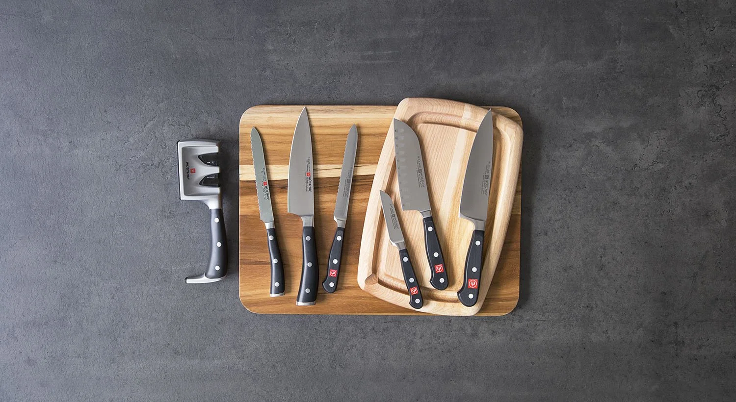 A set of six kitchen knives with black handles is displayed on two wooden cutting boards against a dark gray textured background. The knives vary in size and shape, including a cleaver, and are neatly arranged.