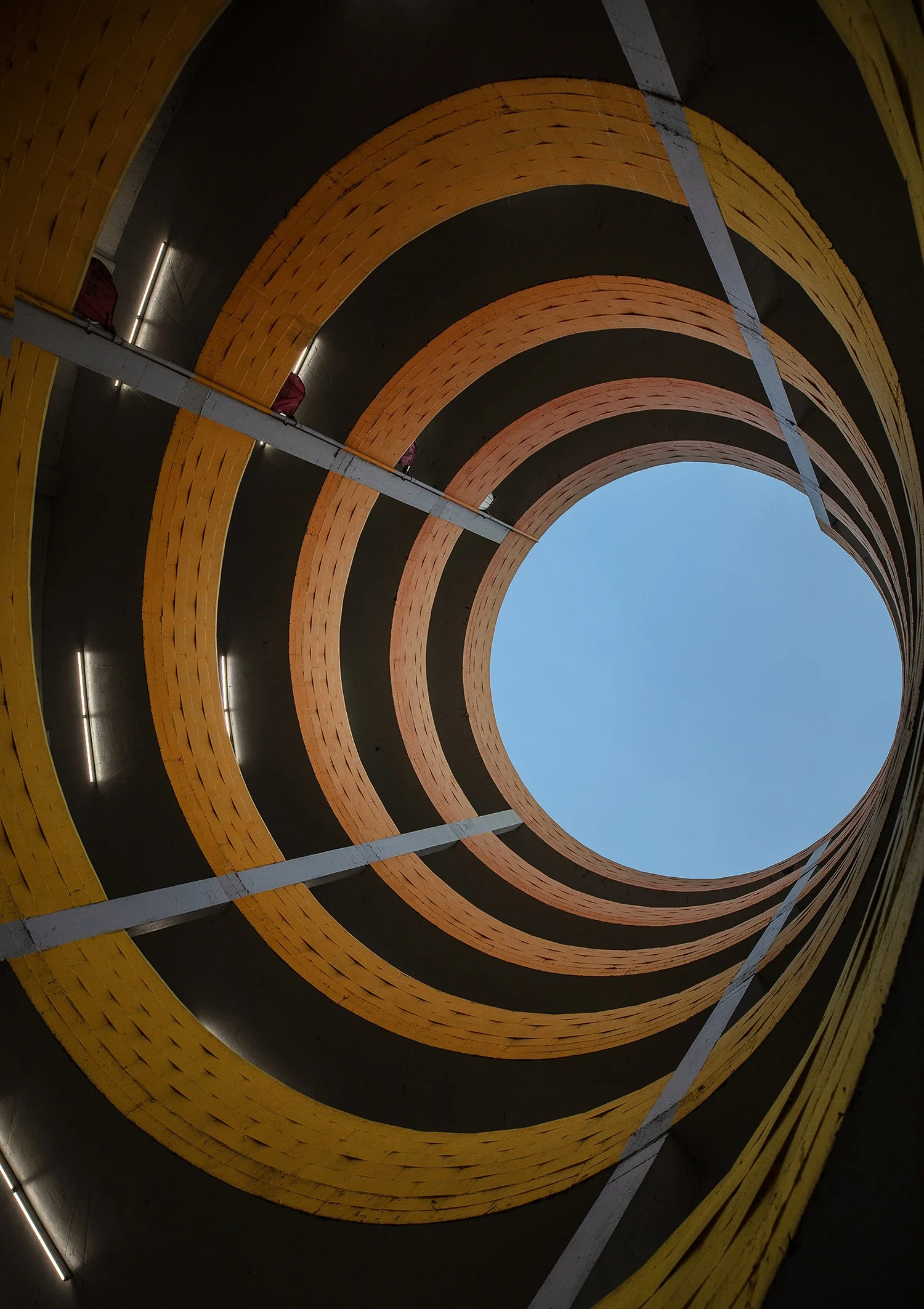 View from the bottom of a cylindrical parking garage, featuring concentric yellow and gray spirals leading up to the sky. The architectural design creates a tunnel-like visual effect.
