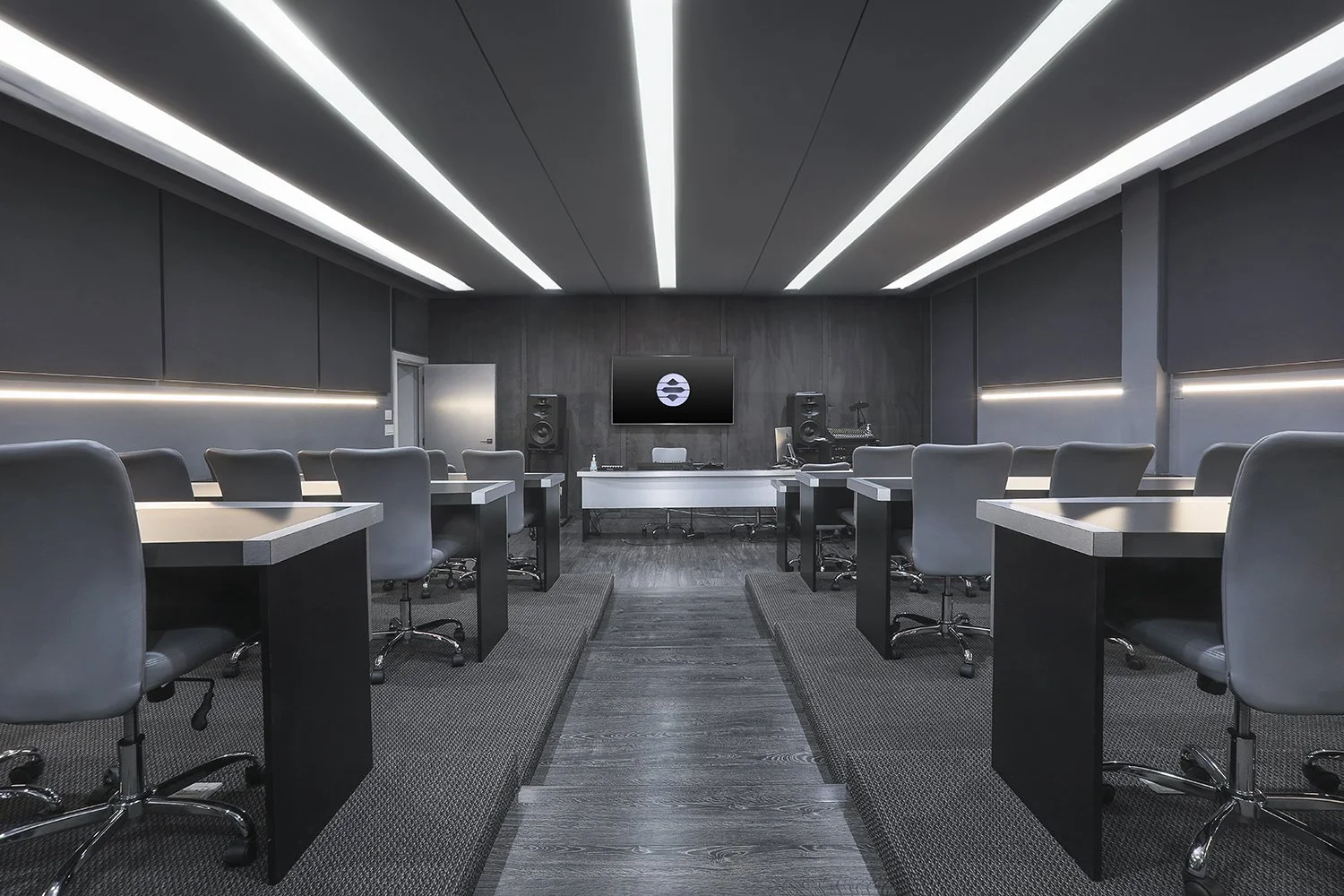 A modern conference room with sleek, minimalist décor. It features a long aisle leading to a large screen mounted on the front wall. Rows of tables with gray chairs line either side of the aisle. The room is illuminated by linear ceiling lights.