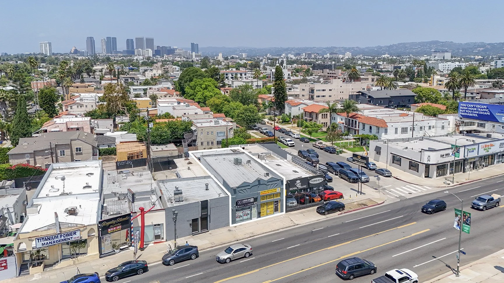 Aerial view of a busy city street lined with small businesses and parked cars, with residential neighborhoods and tall buildings in the background under a clear blue sky.