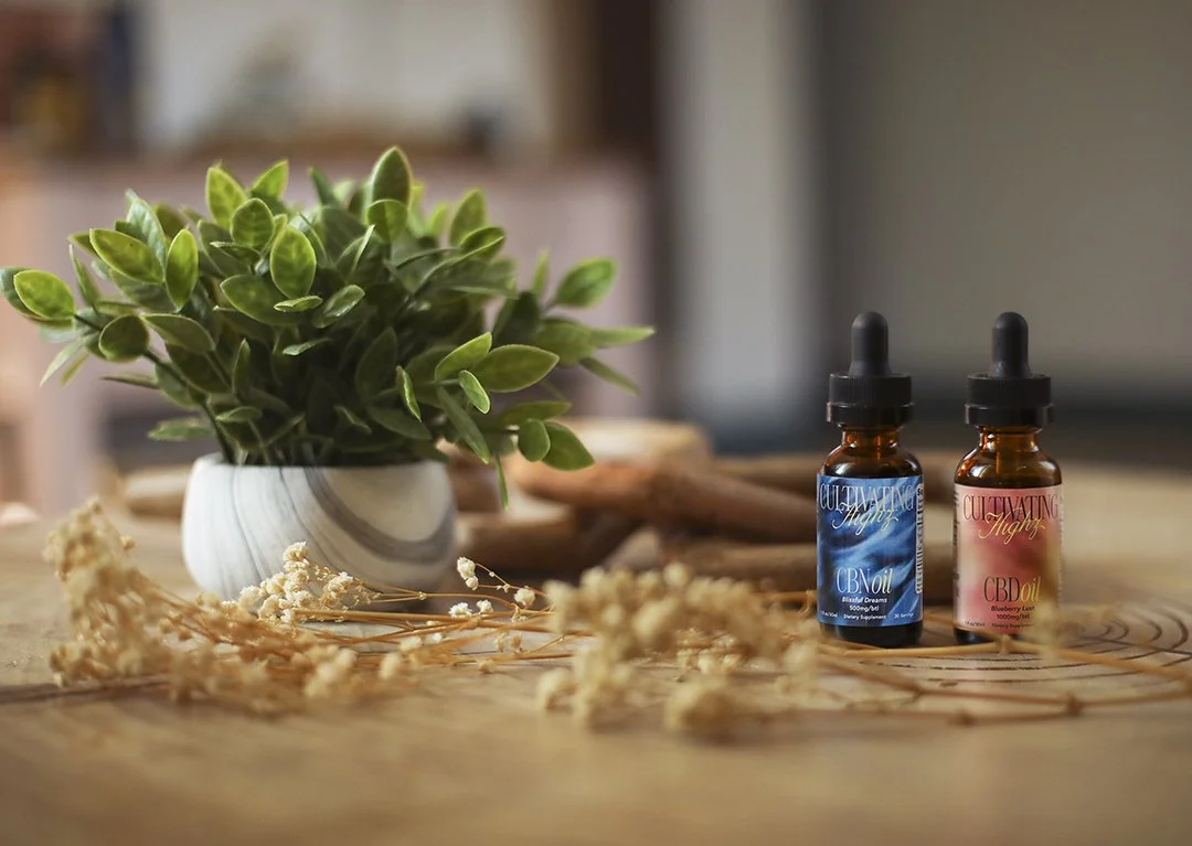 Two bottles with dropper caps, labeled CBD oil, are placed on a wooden table decorated with dried flowers. A small, leafy plant in a marbled pot is nearby, creating a natural and calming atmosphere.