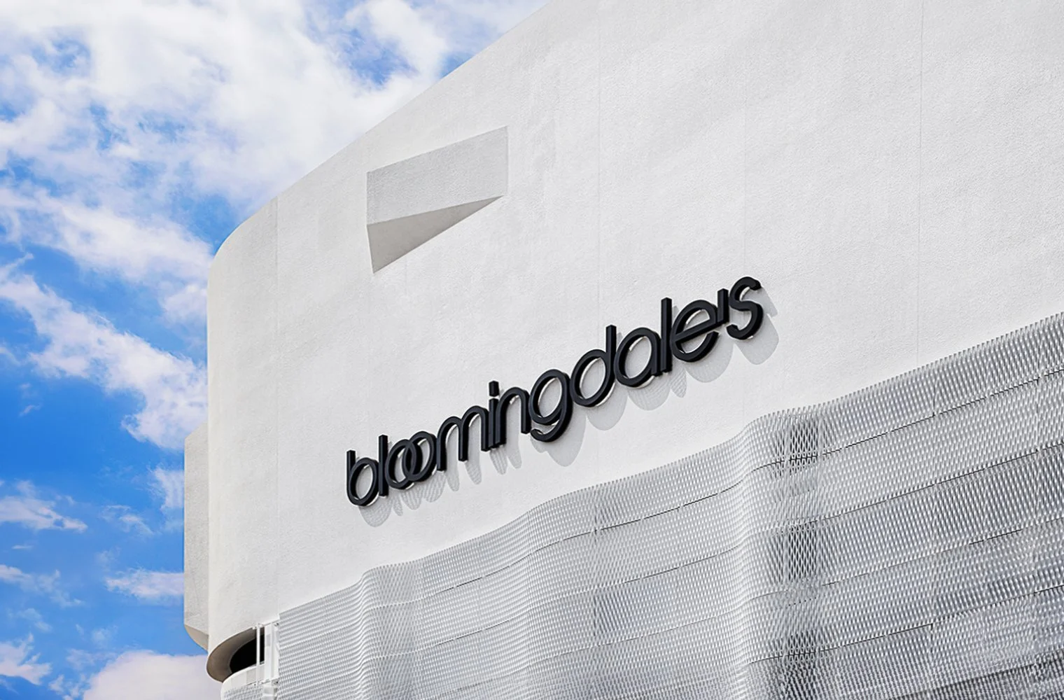The exterior of a modern white building with the Bloomingdale's logo prominently displayed in black letters against the upper part of the wall. The building has a curved structure and mesh-like detailing near the bottom. The sky is clear with a few s
