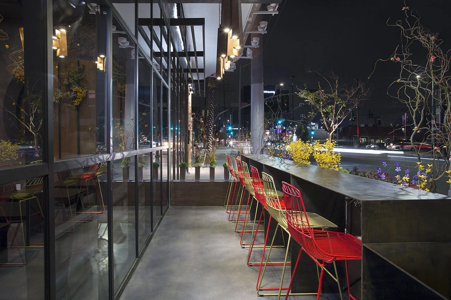 A modern outdoor seating area at AB Steakhouse in Beverly Hills at night with red wire chairs and a long black countertop. The area is adorned with plants and flowers. Large glass windows reflect the vibrant city lights, and the street outside is ill