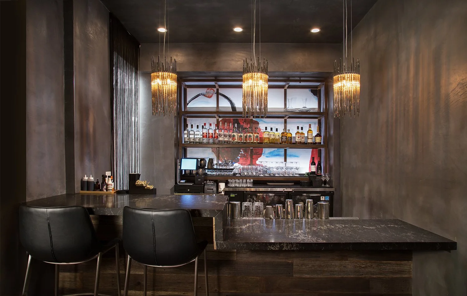 A modern, dimly lit bar with two black bar stools in front of a sleek, dark-colored countertop. Behind the bar are shelves stocked with various bottles of liquor, with three contemporary chandeliers hanging from the ceiling, adding a stylish touch to