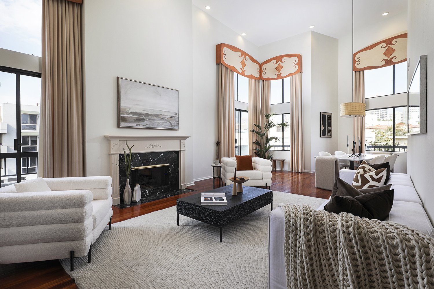 A spacious living room with high ceilings, large windows, and a fireplace. It features white sofas, a black coffee table, and a decorative rug. Tall plants and abstract art complete the elegant look, while natural light fills the space.