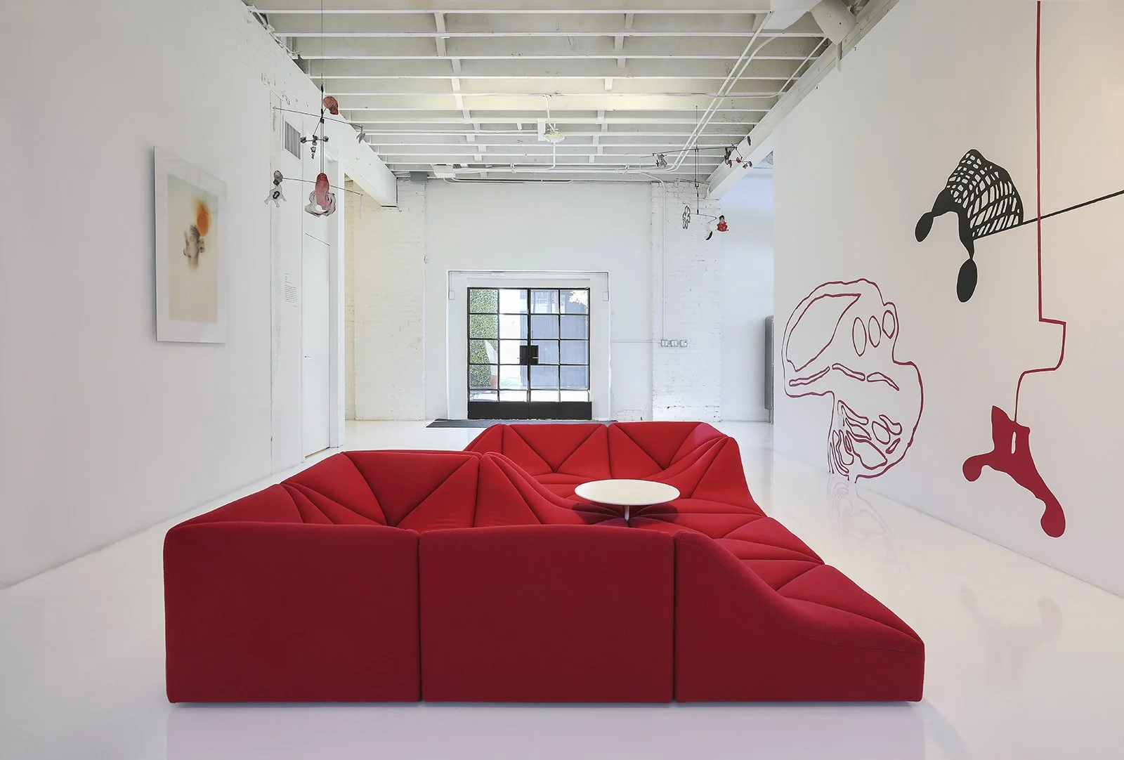 A modern, minimalist room at the Ralph Pucci store in Los Angeles, with a white interior and high ceiling. It features a geometric red sectional sofa with a round white side table. Art pieces hang on the white walls, and a large window at the back le