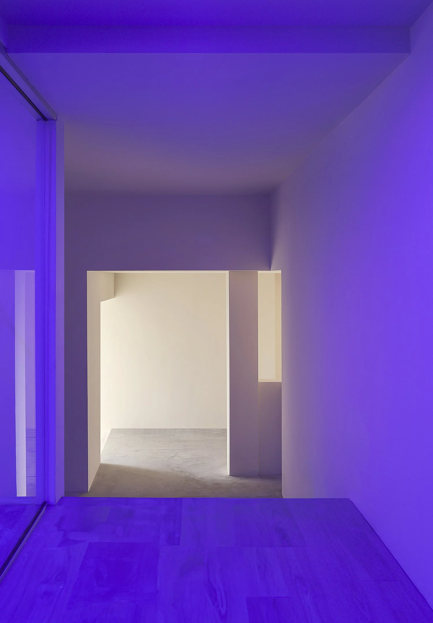 A minimalistic hallway featuring clean lines and smooth surfaces. The walls are illuminated by a soft purple light, while a warm yellow light emanates from a recessed area further down the hall. The floor is made of light-colored wood, enhancing the 