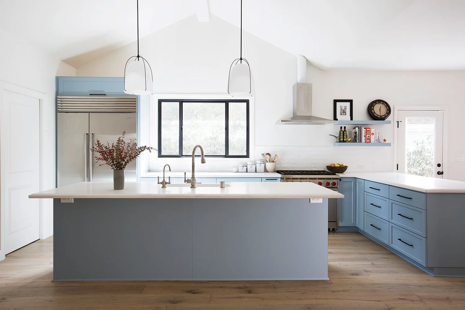 Modern kitchen with light blue cabinets and island, white countertops, and stainless steel appliances. Two pendant lights hang over the island. A window above the sink lets in natural light. Wooden flooring and minimalistic decor complete the aesthet