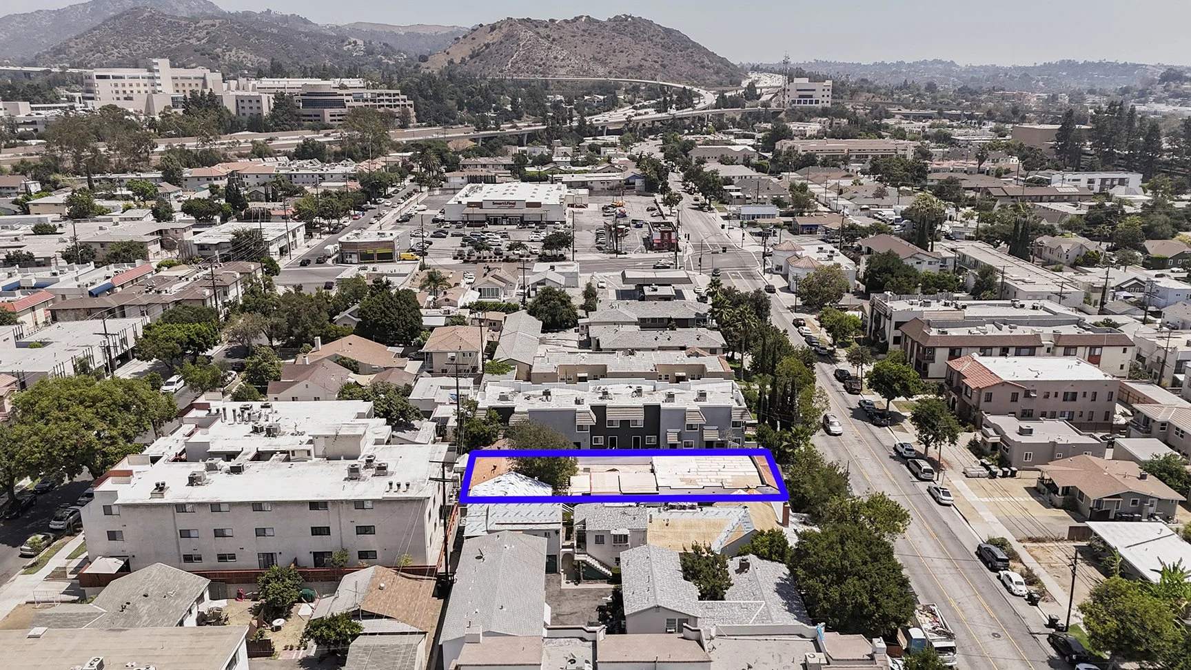 Aerial view of a residential neighborhood with a cluster of buildings; one rectangular property is outlined in blue near the bottom center of the image. Trees line the streets and mountains are visible in the background.