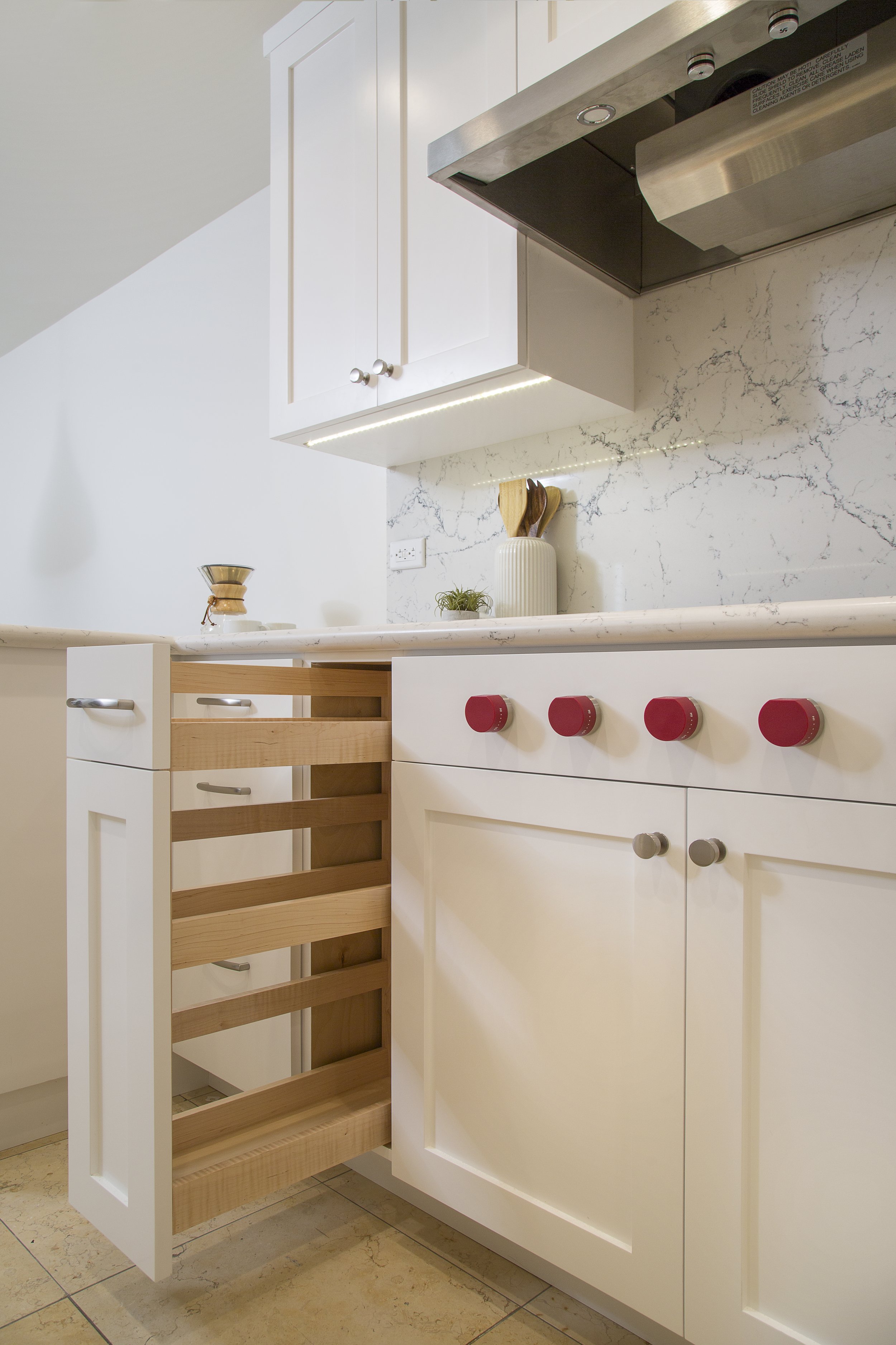 A modern kitchen with white cabinets, red stove knobs, a marble backsplash, and a narrow pull-out wooden spice rack drawer partially open beneath the countertop.