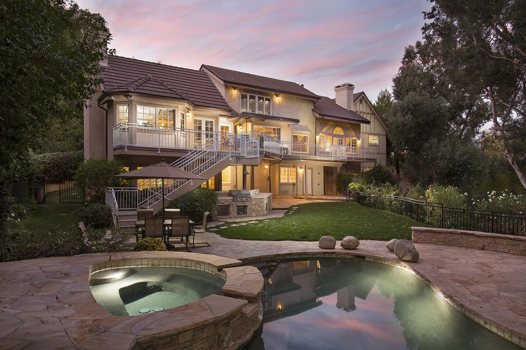 A luxurious two-story house with multiple balconies and large windows overlooks a backyard featuring a landscaped garden, stone patio, outdoor seating area, built-in grill, and a stone-edged pool with an integrated hot tub. The sky shows a picturesqu