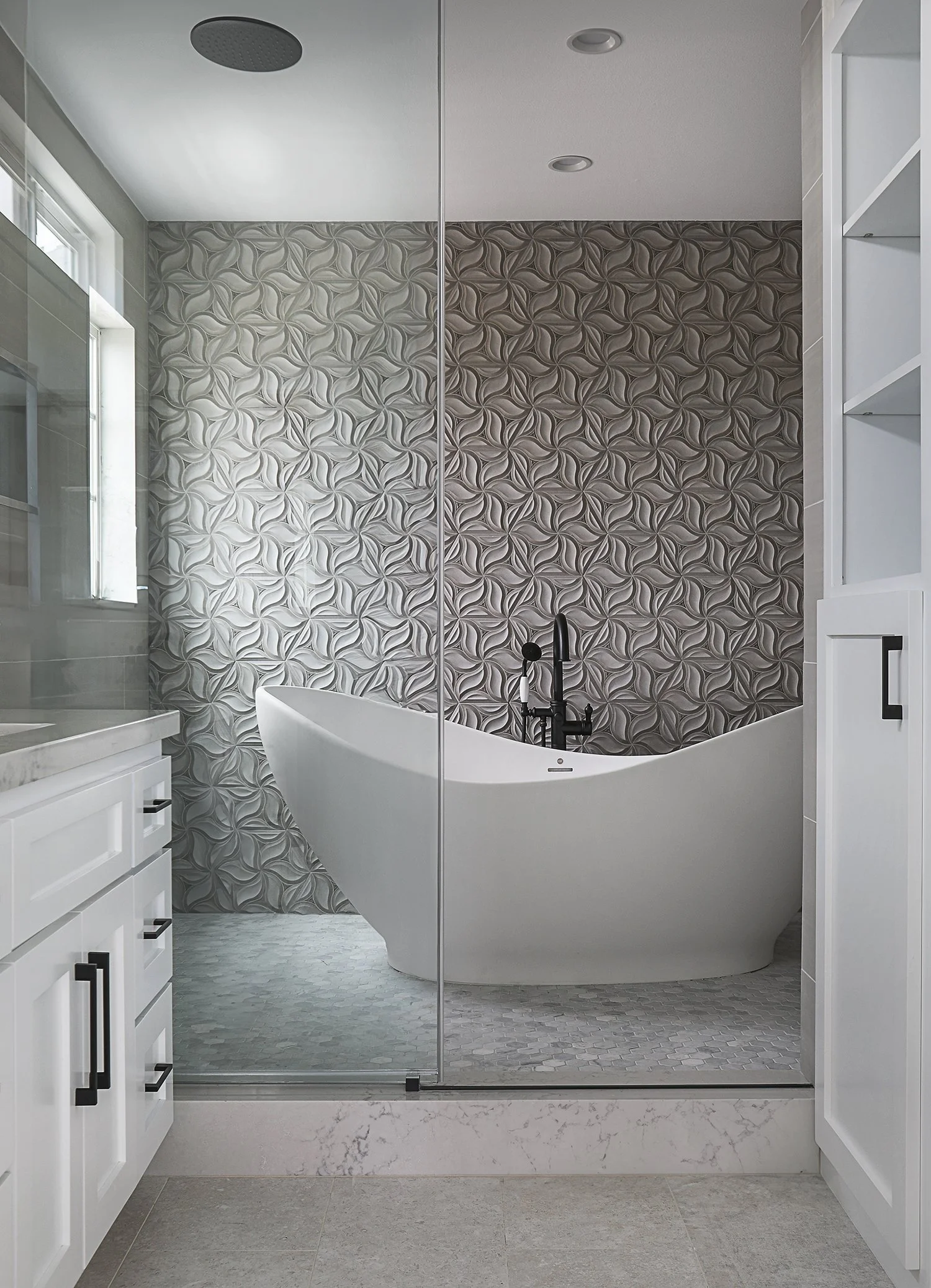 A modern bathroom featuring a white freestanding bathtub against a wall with geometric patterned tiles. A glass shower enclosure is to one side, and built-in white cabinets with black handles are on the other. Light filters in through a window, illum