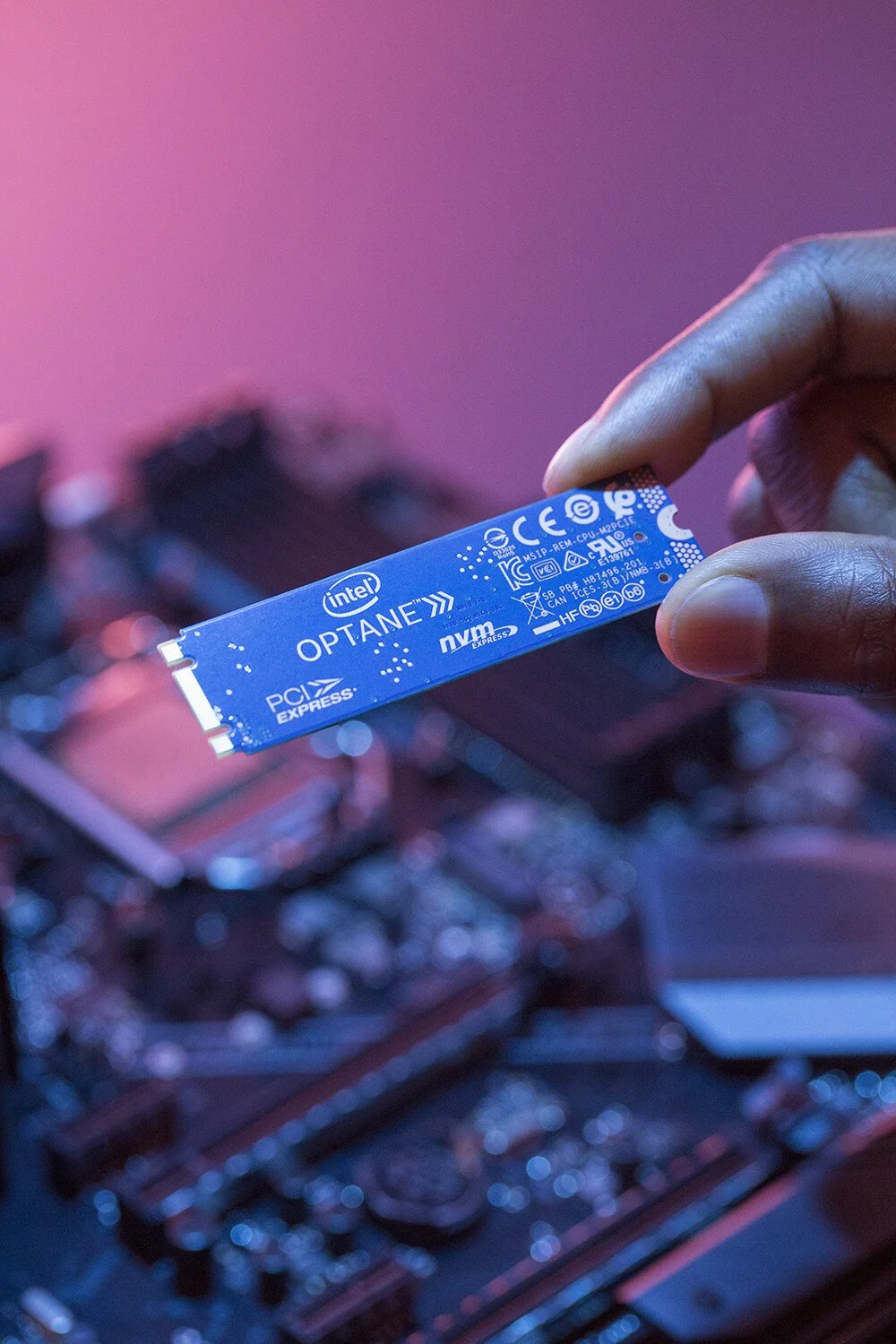 Close-up of a hand holding an Intel Optane NVMe SSD against a blurred background of computer components. The SSD is blue with white text and symbols, highlighting its compact design. The lighting is a blend of pink and blue tones.