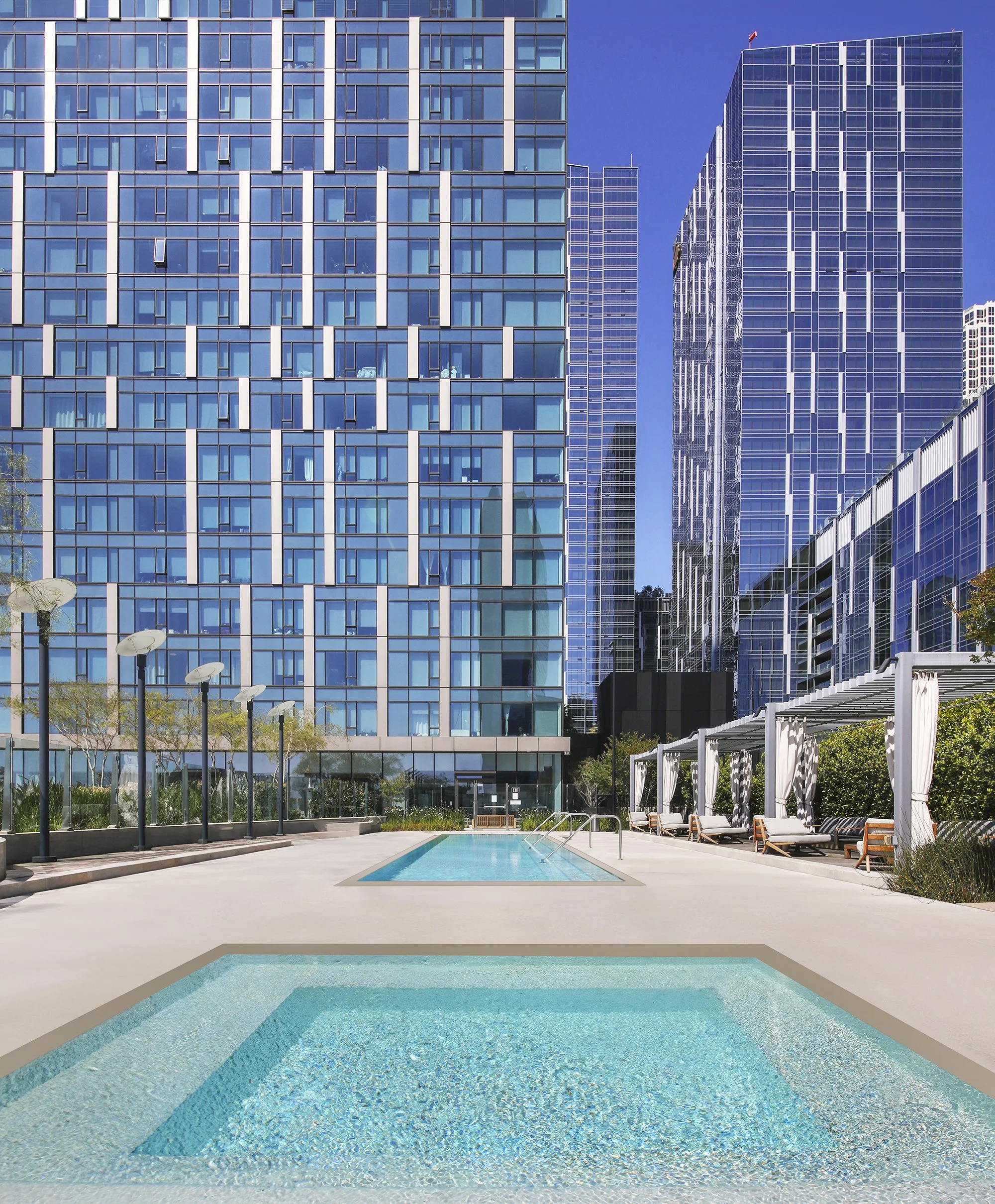 Modern urban outdoor pool area surrounded by tall, glass-covered skyscrapers and apartment buildings. The clean, rectangular pools reflect the blue sky. Several white cabanas and lounge chairs line one side of the pool deck, creating a relaxing atmos