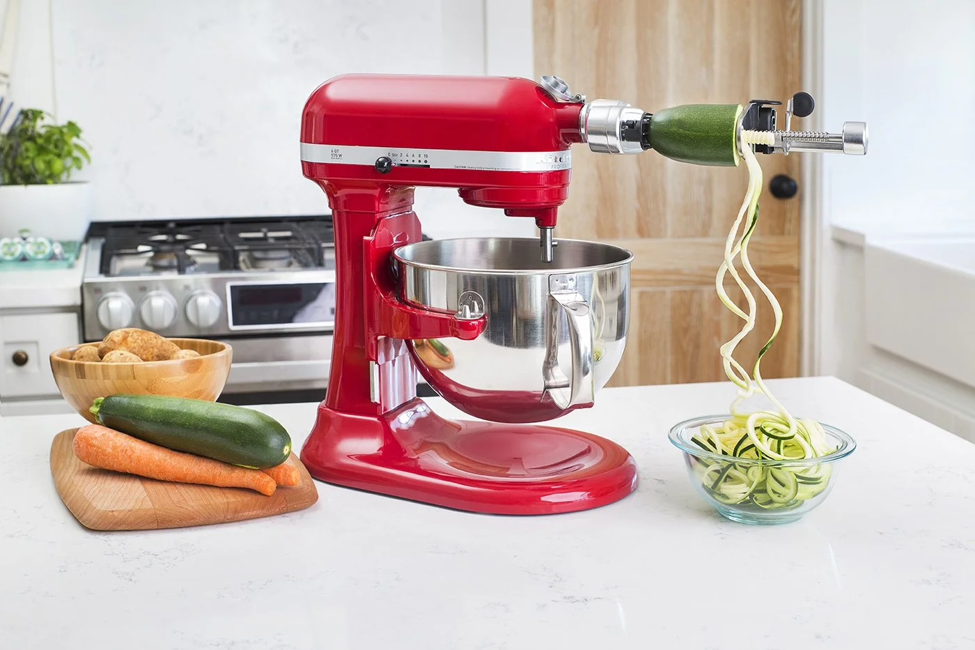 A red stand mixer with a spiralizer attachment is on a kitchen counter, creating zucchini noodles into a glass bowl. Nearby are a cutting board with a carrot and zucchini, and a bowl of potatoes. A stove is in the background.