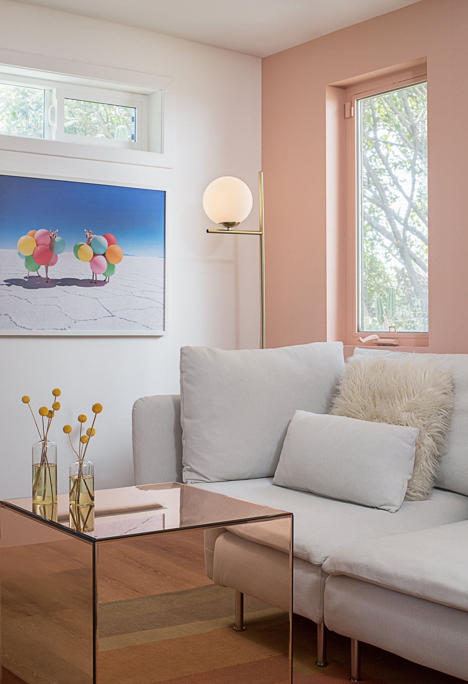 A cozy living room features a white sectional sofa with fluffy pillows, a modern glass coffee table, and a gold floor lamp with a globe bulb. The walls are painted in soft pink and white, with a large window and framed artwork of colorful birds.