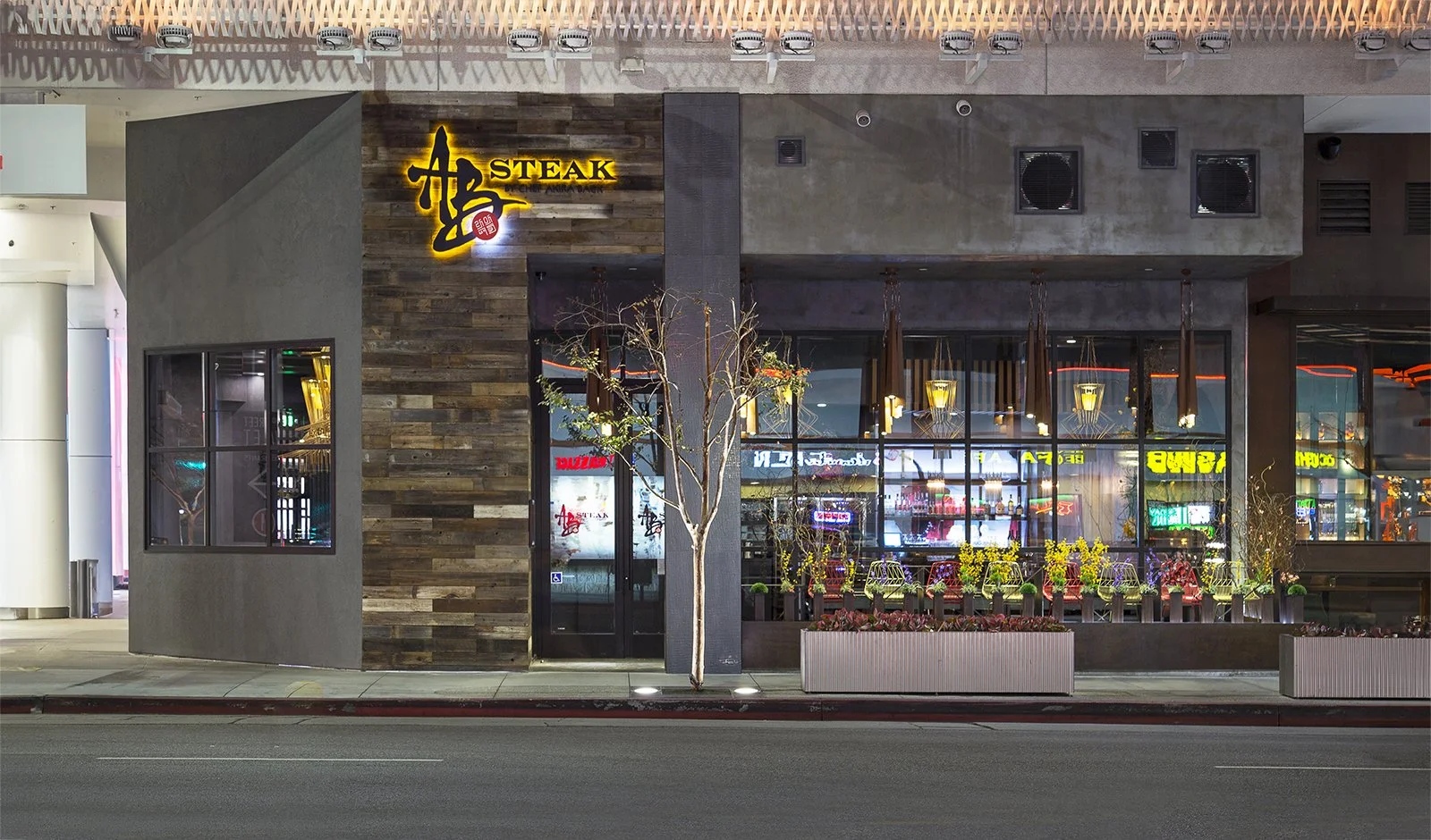 AB steakhouse restaurant exterior at night with a modern design, featuring a mix of concrete and wooden textures. The illuminated yellow sign reads "Steak," and the entrance is adorned with plants. The large windows reveal a warmly lit interior with 