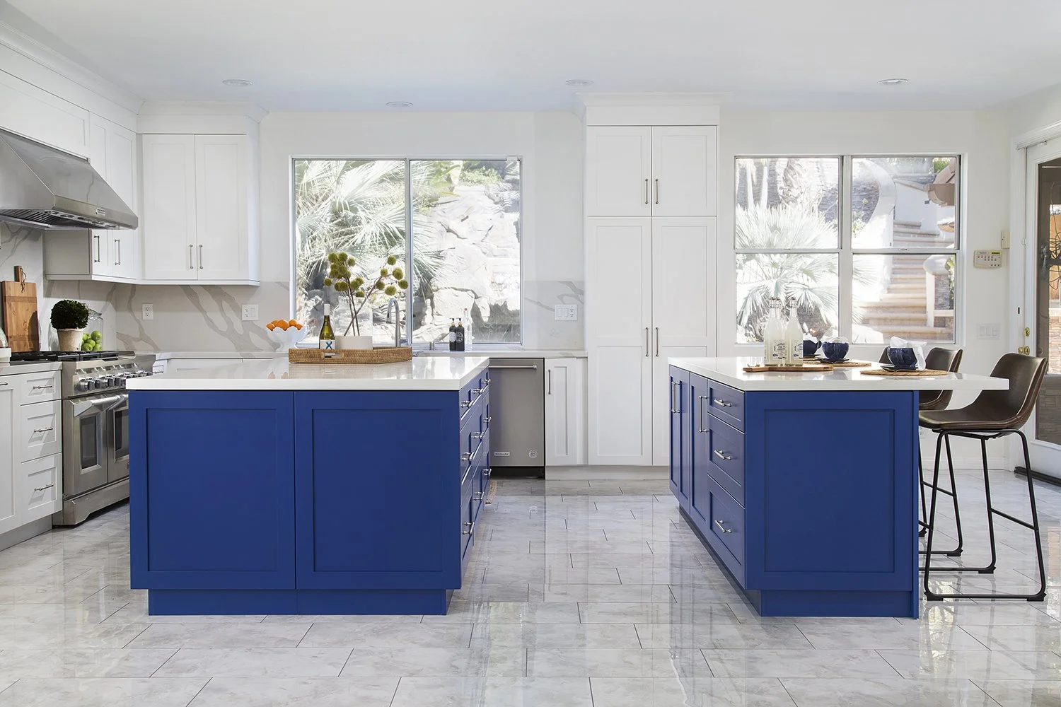 A modern kitchen featuring two islands with blue cabinets and white countertops. The space is brightened by large windows, with visible stainless steel appliances and bar stools. The floor is a light-colored polished tile.