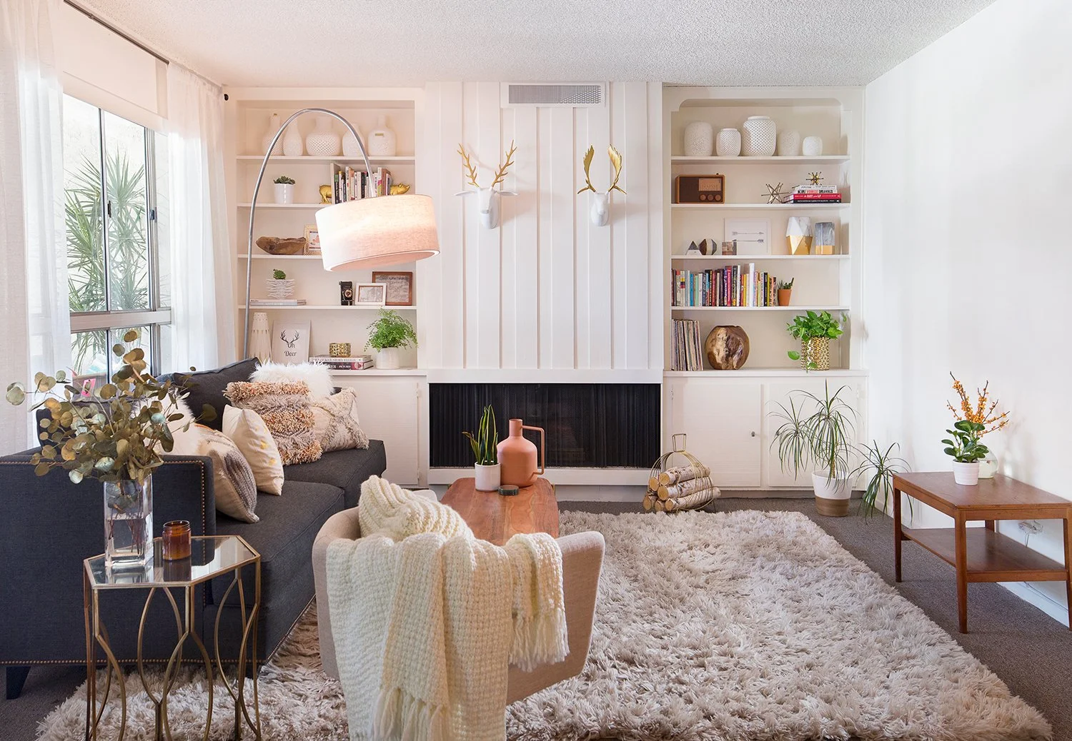 A cozy living room with a modern design features a gray sectional sofa with plush cushions, a wooden coffee table, and a fluffy white rug. Built-in shelves with books and decor flank a central fireplace. A tall floor lamp arcs over the sofa, and larg