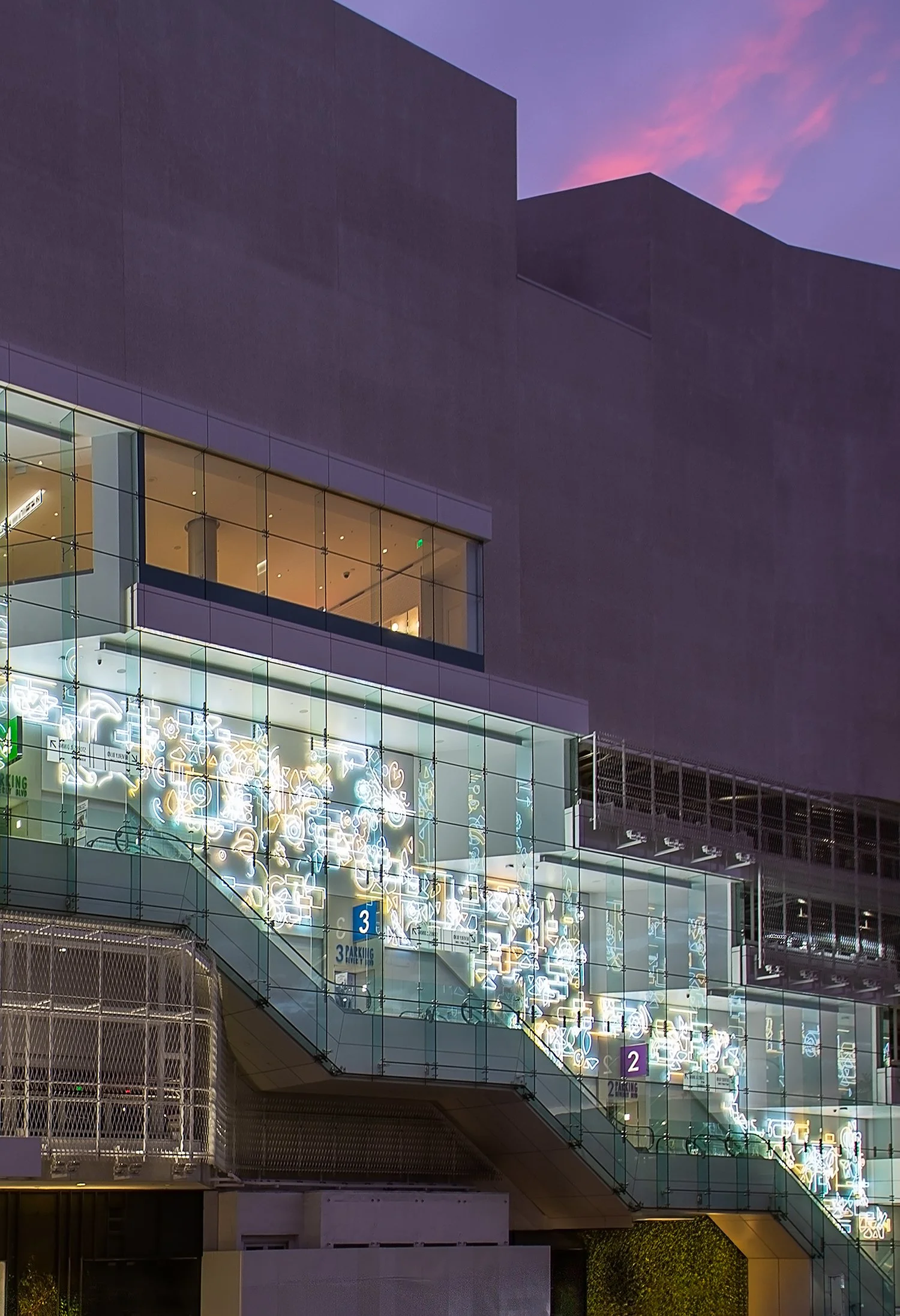 A modern building at twilight with reflective glass panels, showcasing an entrance with a large staircase. Several floors are visible through the glass, and some lights are on inside. The exterior architecture features angular shapes and purple light