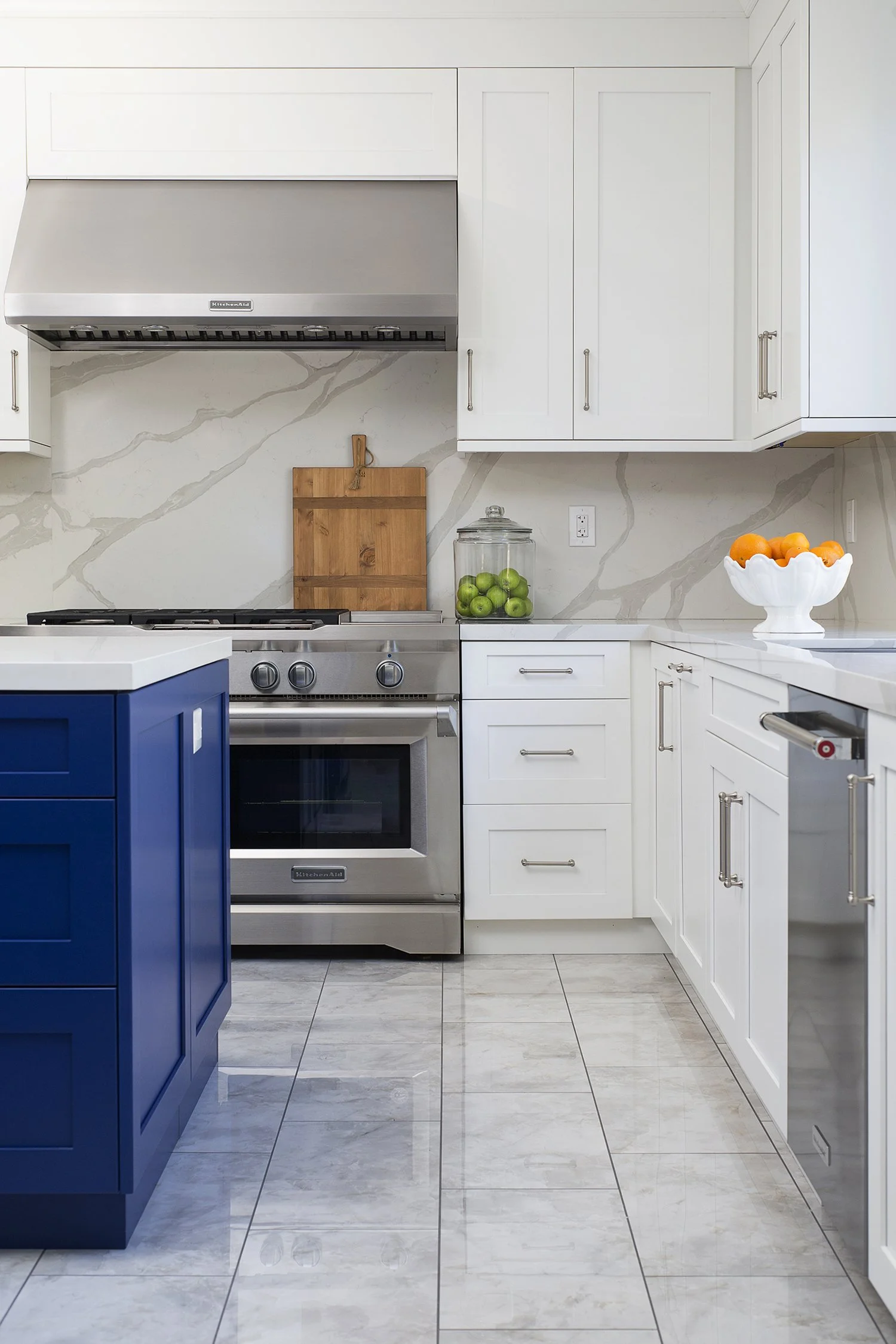 Modern kitchen with white cabinets and stainless-steel appliances. A large blue island with a quartz countertop stands centrally. On the counter, there's a white bowl with oranges. The backsplash features a marble-like pattern, and a jar with green a