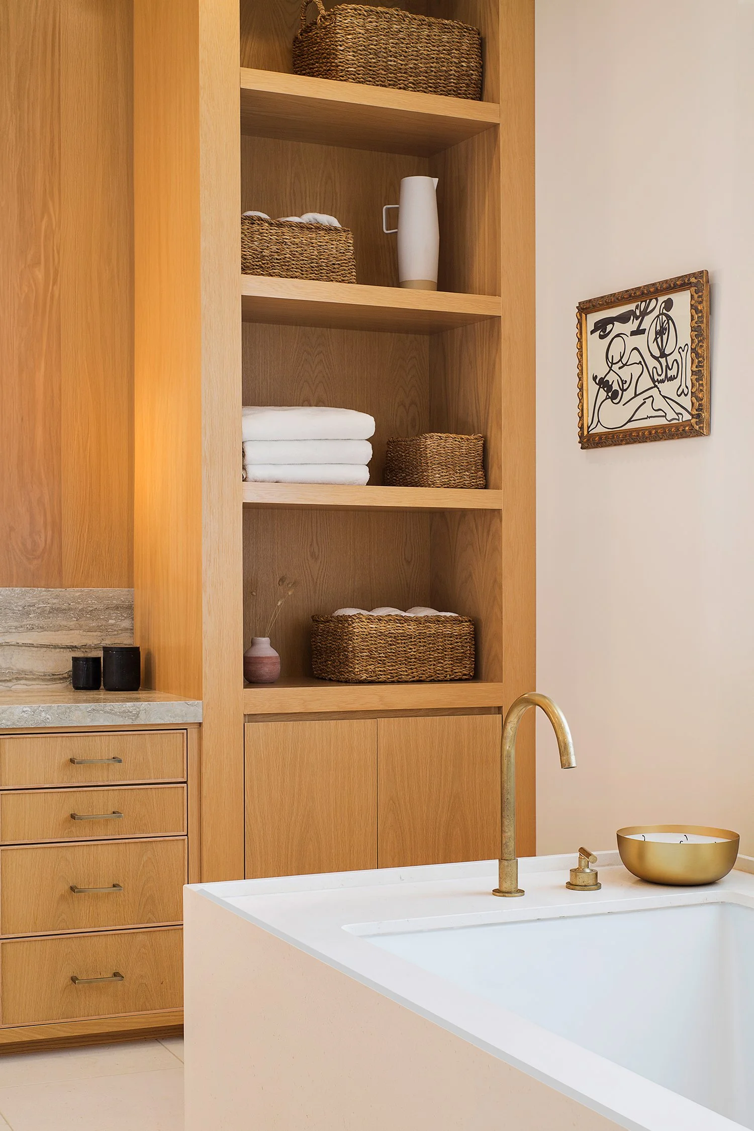 A modern bathroom with a white bathtub featuring a gold faucet. Adjacent, wooden shelves hold wicker baskets, white towels, and decor items. A wooden cabinet with drawers is topped with a marble surface. A framed abstract artwork hangs on the light-c