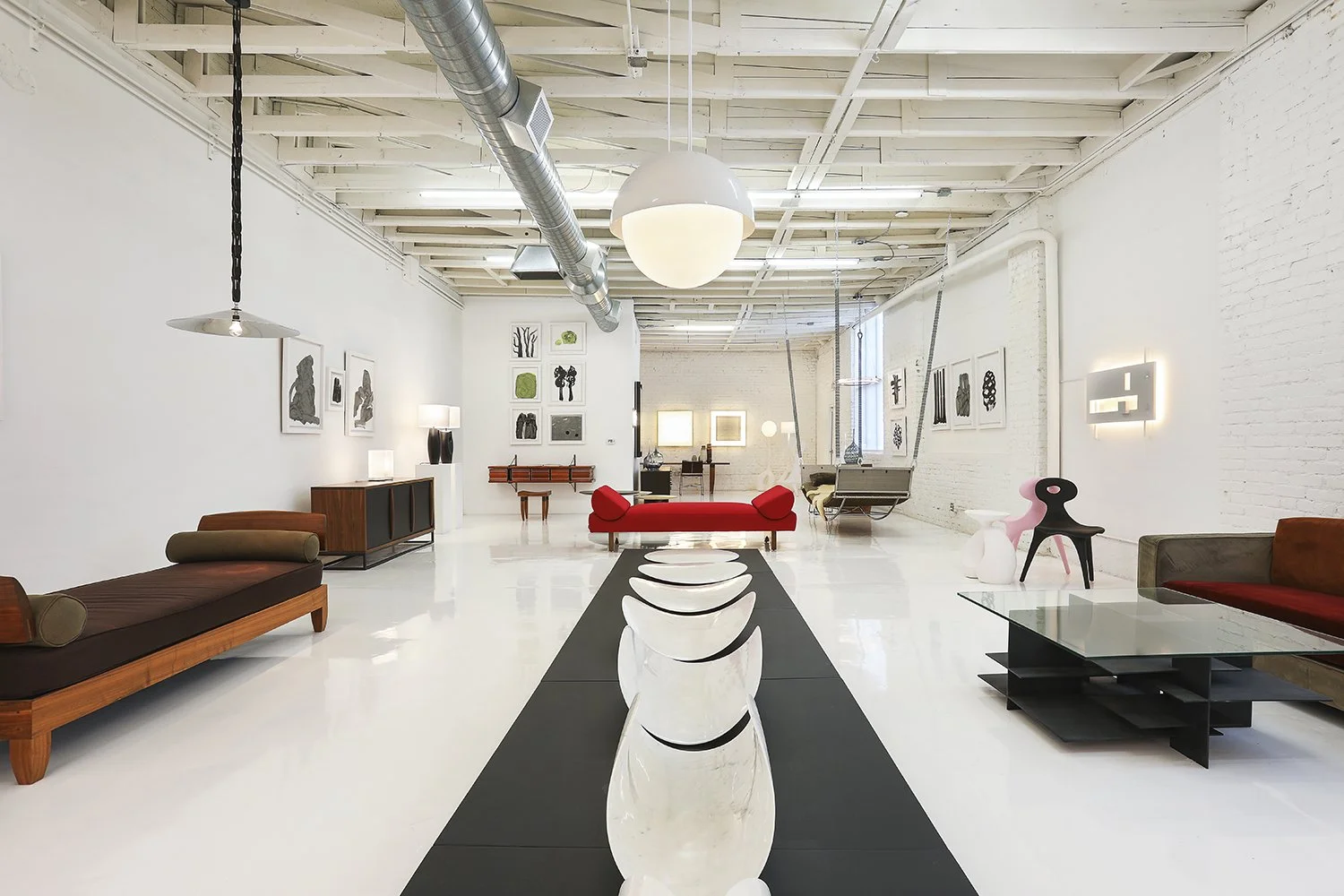 A modern loft space with white walls and flooring, featuring various contemporary furniture pieces. The space has an industrial design with exposed beams and ductwork. The decoration includes abstract art on the walls and a minimalist arrangement of 