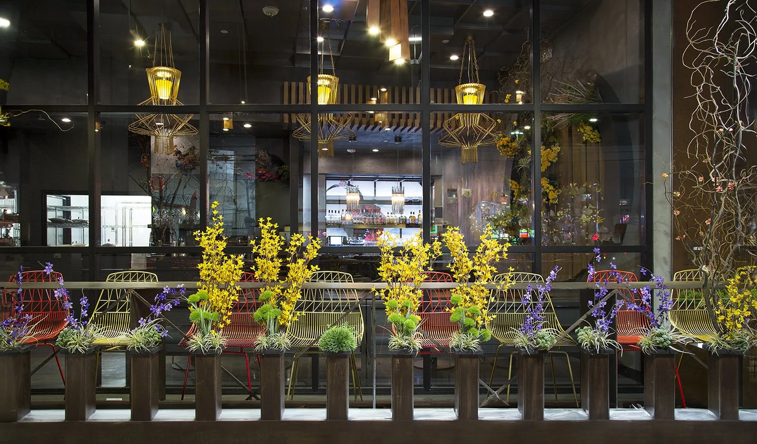 A chic, modern café exterior of AB Steakhouse restaurant, featuring a row of colorful metal chairs paired with a communal high table adorned with bright yellow and purple flowers. The background showcases an illuminated counter area with various deco