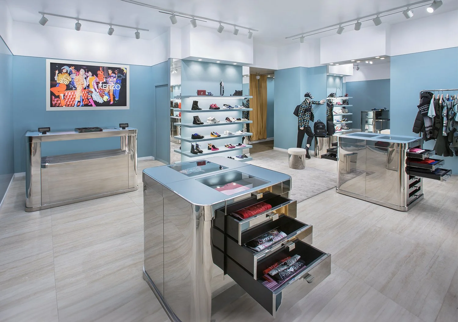 Zara's retail store interior with sleek metallic and glass displays. The store features light blue walls, recessed lighting, and wooden flooring. Shelves and tables display neatly organized clothing, shoes, and accessories. A digital screen showcases