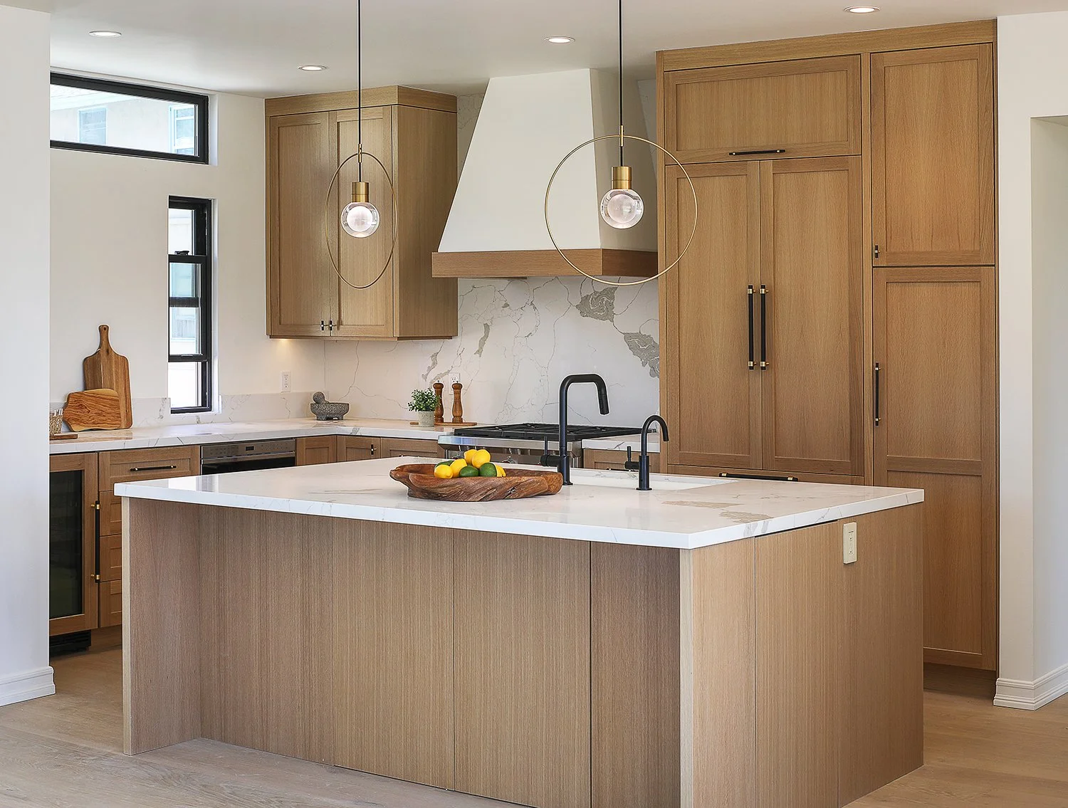 A modern kitchen with light wood cabinetry, a white marble countertop island, and black fixtures. Two pendant lights hang over the island, which has a wooden fruit bowl. A white hood and marble backsplash are above the stove. Natural light enters thr
