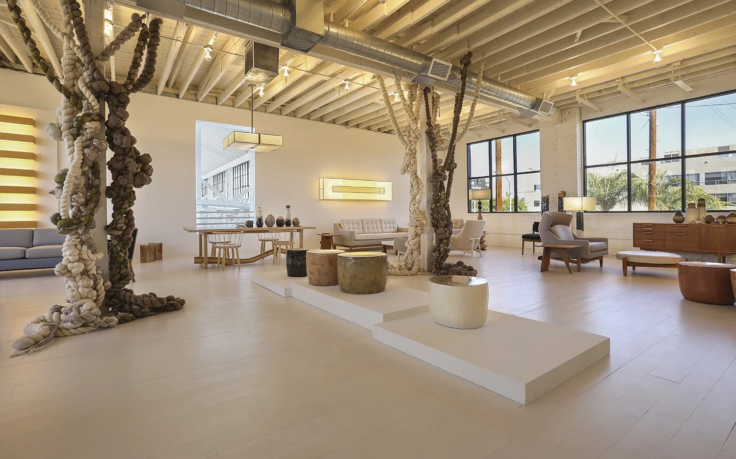 Spacious, minimalist room at the Ralph Pucci store located in Los Angeles. It has large windows, light wooden floors, and exposed ceiling beams. The room is decorated with modern furniture, including seating areas, tables, and several potted plants. 