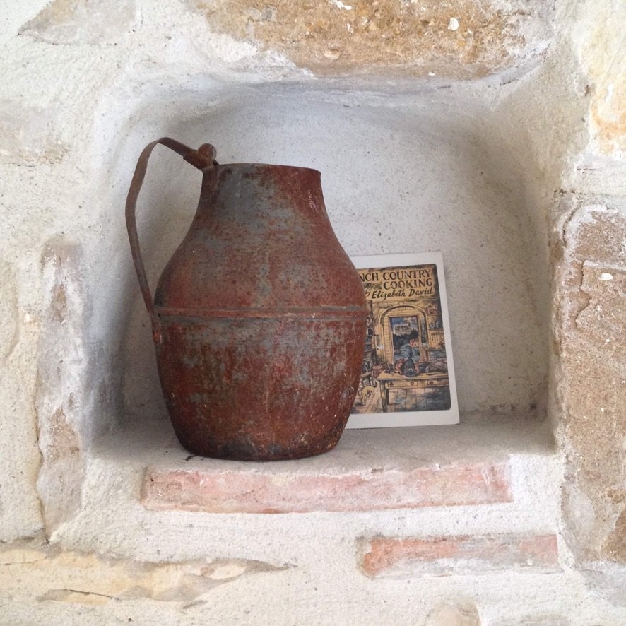 A rustic metal jug in a stone niche in front of a card depicting Elizabeth David's book French Country Cooking