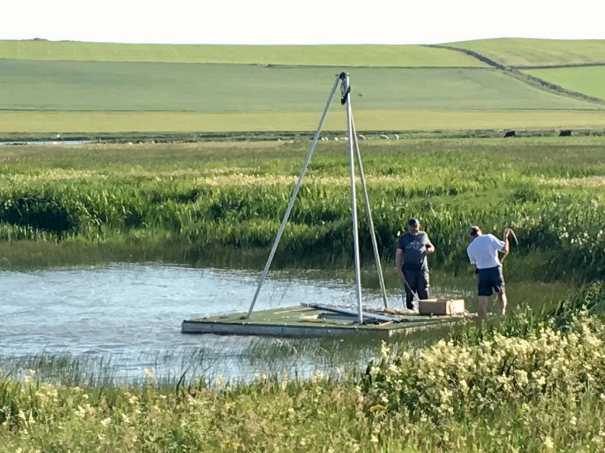 Archaeologists preparing a raft to take cores in the loch of Skaill. This picture also illustrates how different Skara Brae is from the era depicted in The Boy With The Bronze Axe . The picture is taken by Sue Dawson and the work is funded by Historic Environment Scotland.