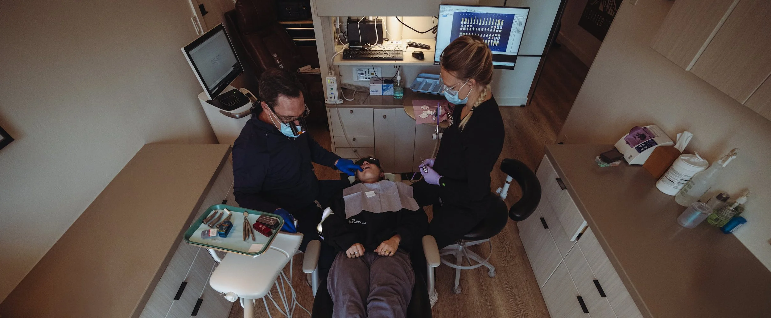 Dentist or dental technician examining a patient's teeth while seated in a dental chair, with dental tools and equipment nearby.