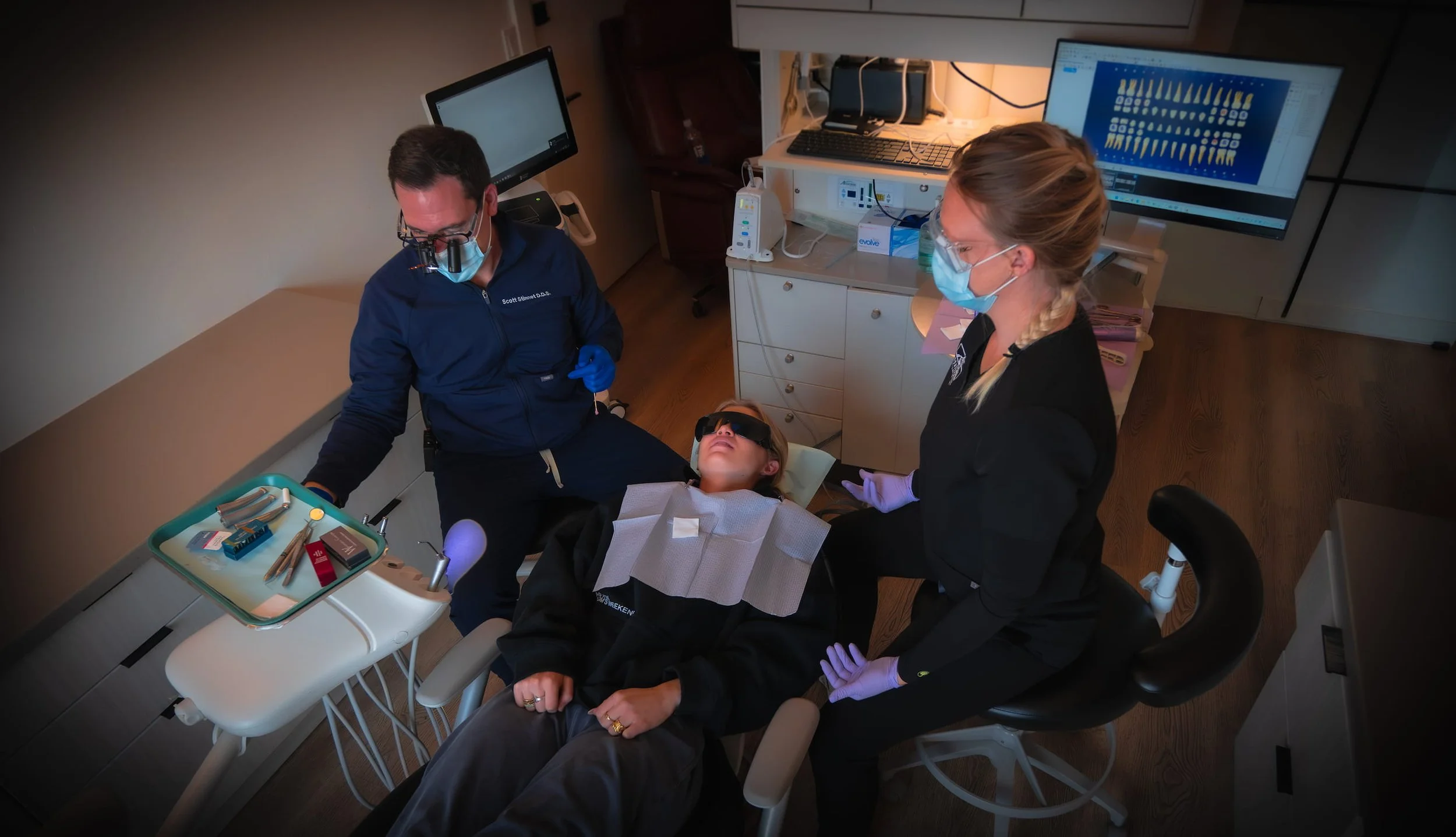 A patient wearing protective eyewear and a dental bib lying on a dental chair in a dental clinic, flanked by two dental professionals wearing masks and gloves, with dental tools and equipment nearby.